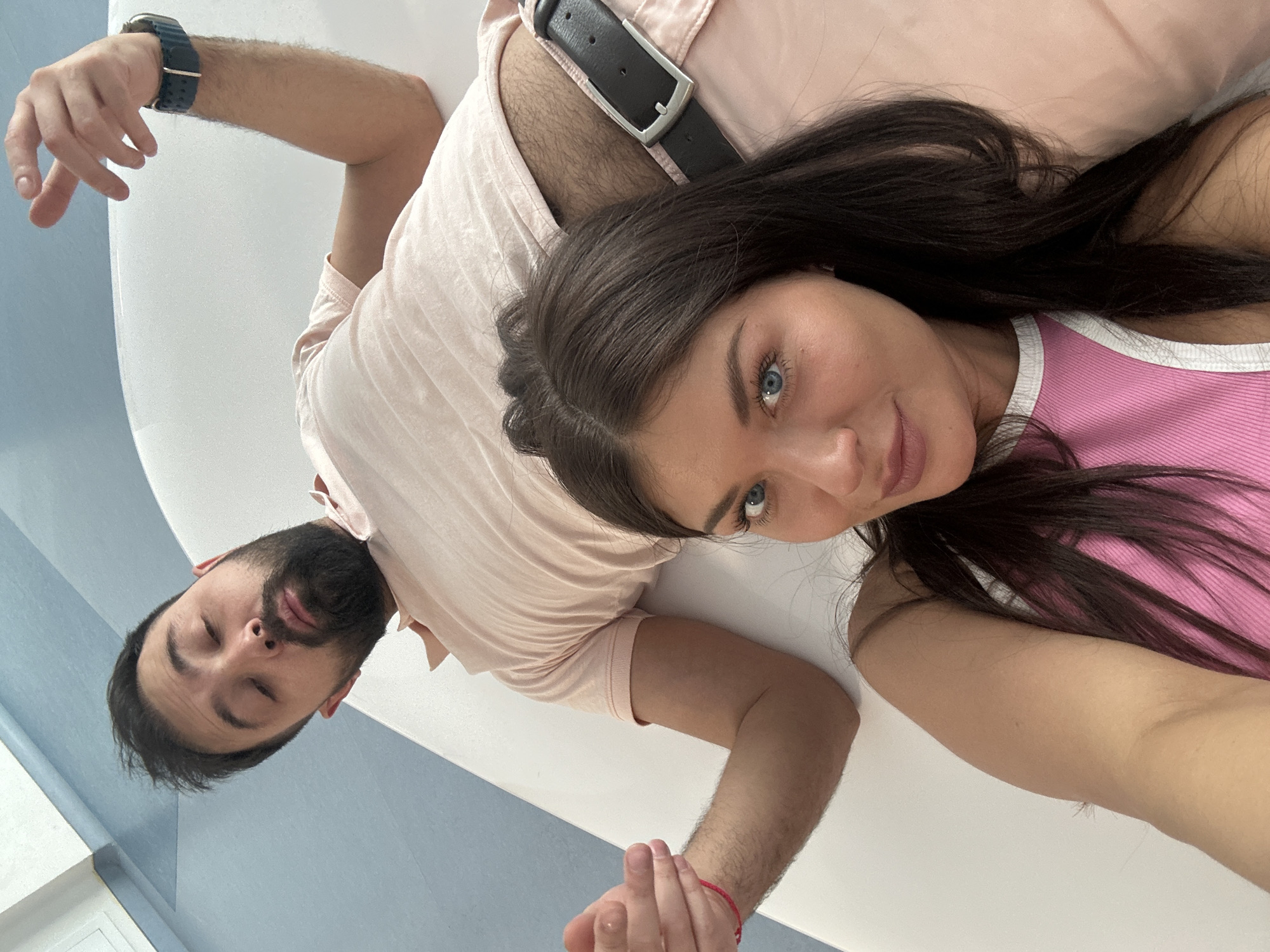 A man and woman are lying down side-by-side on a surface, possibly a floor. The woman is on the right side of the image and appears to be taking a selfie. She has dark hair and blue eyes, wearing a pink top with a white trim. The man has a beard and dark hair, wearing a pink shirt. He is lying on his back with his eyes closed. Both are wearing pink bottoms. The man is wearing a watch. A black leather belt is visible. The background appears to be a white and blue surface. The city location is not visible in the image.