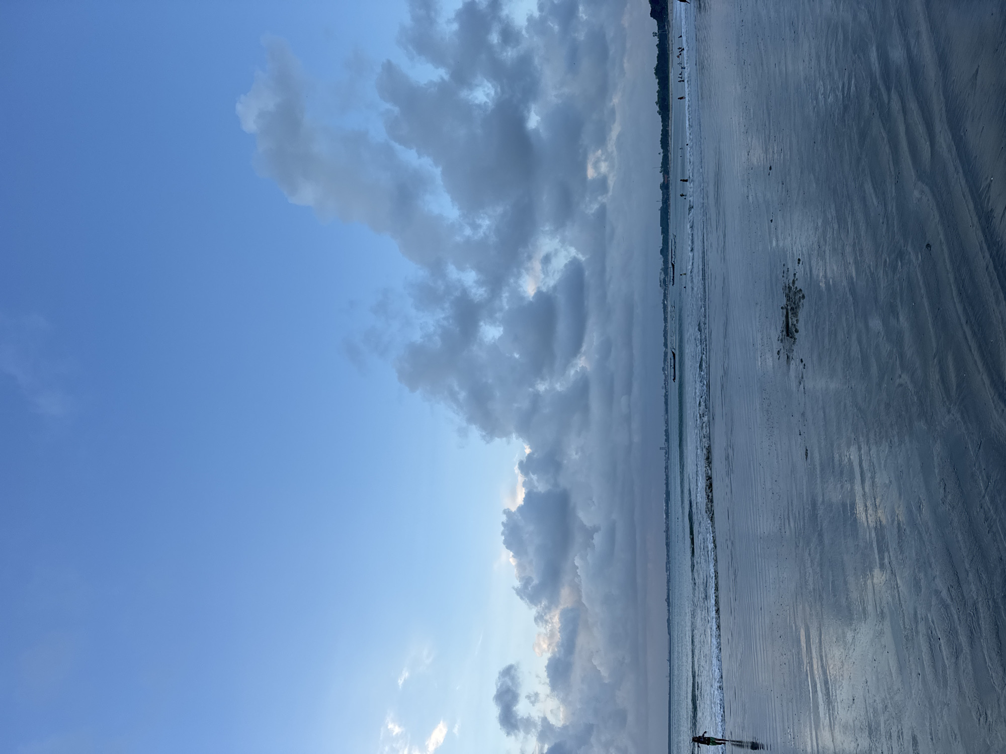 This image captures a serene beach scene, likely in Kuta Selatan, Indonesia, during the late afternoon or early evening. The horizon line, stretching horizontally across the middle of the frame, separates the sky from the expansive beach and ocean.
The sky occupies the upper half, featuring a blend of clear, soft blue and a broad band of voluminous, greyish-white cumulus clouds. Some parts of the clouds appear brighter, suggesting the low angle of the sun, possibly indicating sunset or pre-sunset hours.
Below the sky, the calm ocean meets a wide, wet sandy beach. Gentle, rhythmic waves create foamy lines along the water's edge. The wet sand is highly reflective, mirroring the sky and clouds, and displays subtle ripples and textures.
Several small, silhouetted figures are visible in the distance along the waterline and on the wet sand, suggesting people are strolling or relaxing. One more prominent figure is seen walking near the water's edge in the foreground. A few small, dark boats or canoes are also scattered along the distant shore. On the far right, a faint, dark landmass, possibly a forested coast or distant town, lines the horizon. The overall atmosphere is peaceful and tranquil. No text is visible in the image.