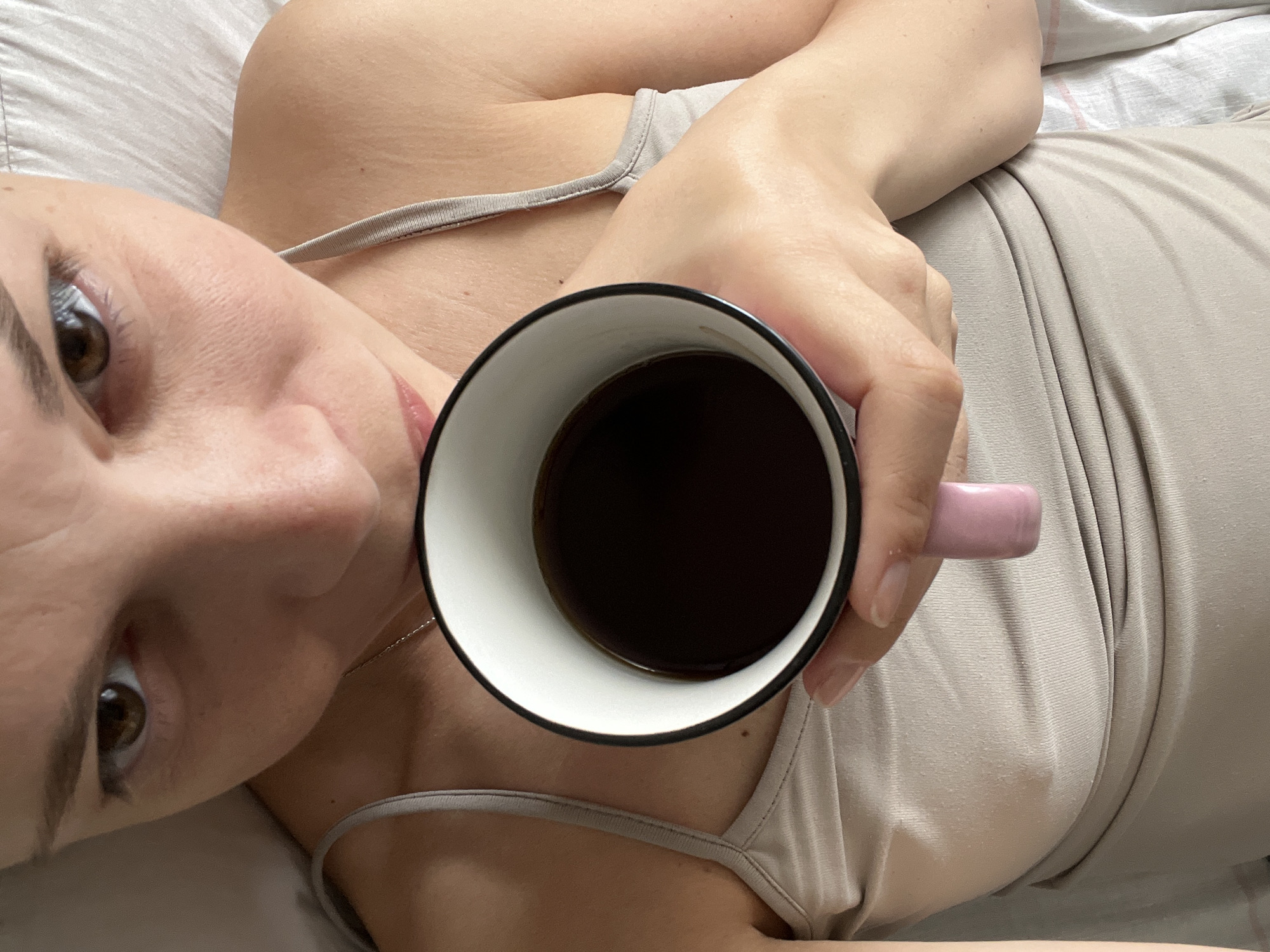 A close-up overhead shot shows a person holding a white mug with a black rim filled with dark liquid, presumably coffee. The person is wearing a light-colored tank top and a silver necklace. Their face is partially visible, with eyes looking towards the mug. The mug has a pink handle. The background consists of white and light-colored fabric, suggesting a bed. The scene is indoors. There is no identifiable information about the city of Tolyatti, Russia.