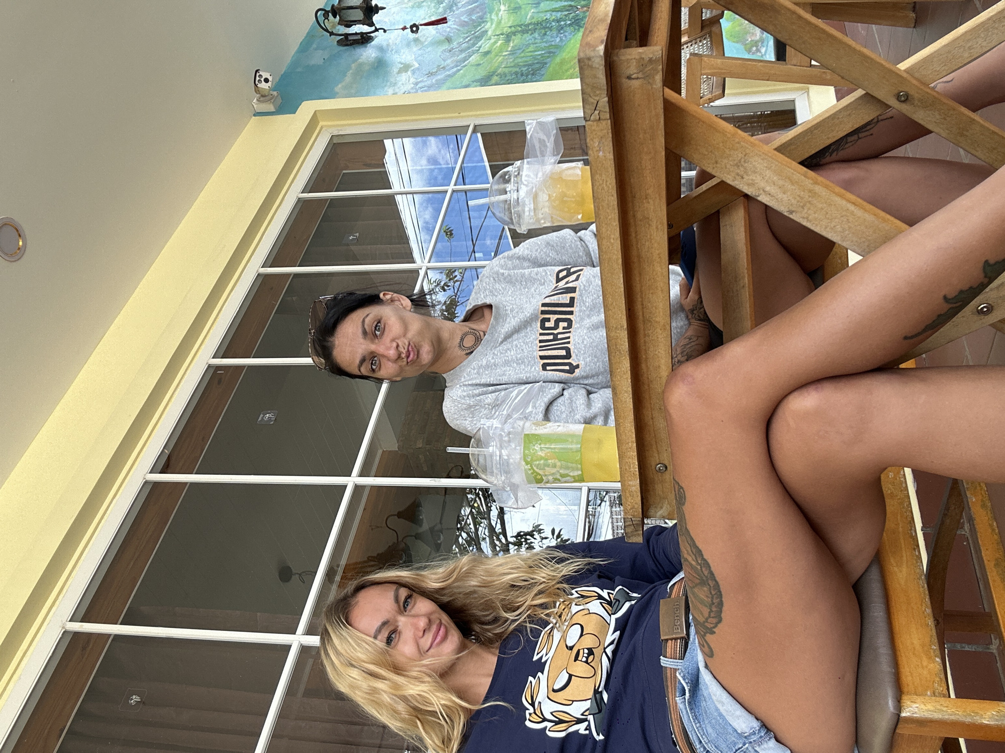 Two women are sitting at a wooden table outdoors. The woman on the left has blonde hair and is wearing a navy blue long-sleeved shirt with a cartoon character graphic and denim shorts. She is looking at the camera and smiling. The woman on the right has dark hair and is wearing a grey sweatshirt. She is looking at the camera with a pursed-lip expression. Both women have drinks in plastic cups with straws in front of them. The background features a large window with a view of trees and buildings, and a mural of a mountain landscape. A security camera is visible on the wall. This scene appears to be in Da Lat, Vietnam, likely at an outdoor cafe.