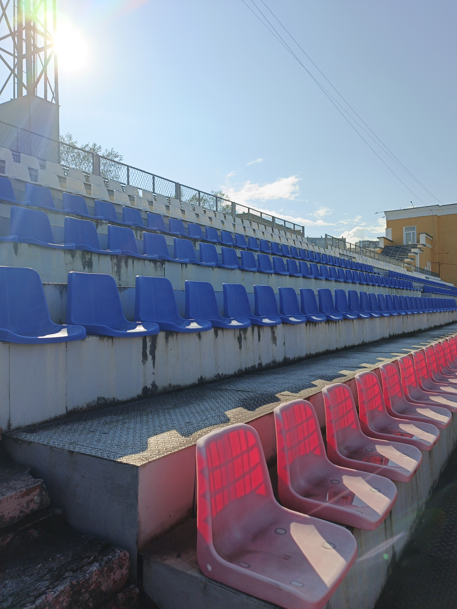 An outdoor stadium with tiered seating. The stadium appears to be located in Zlatoust, Russia, based on the provided context. The seats are arranged in rows, with the lower rows being red and the upper rows alternating between blue and white. The stadium is empty, with no people visible. The sun is shining brightly, casting long shadows on the seats and the ground. The sky is clear blue with a few scattered clouds. There is a metal structure on the left side of the image, possibly a lighting tower. In the background, there are buildings and trees. The overall impression is one of a quiet, empty stadium on a sunny day.