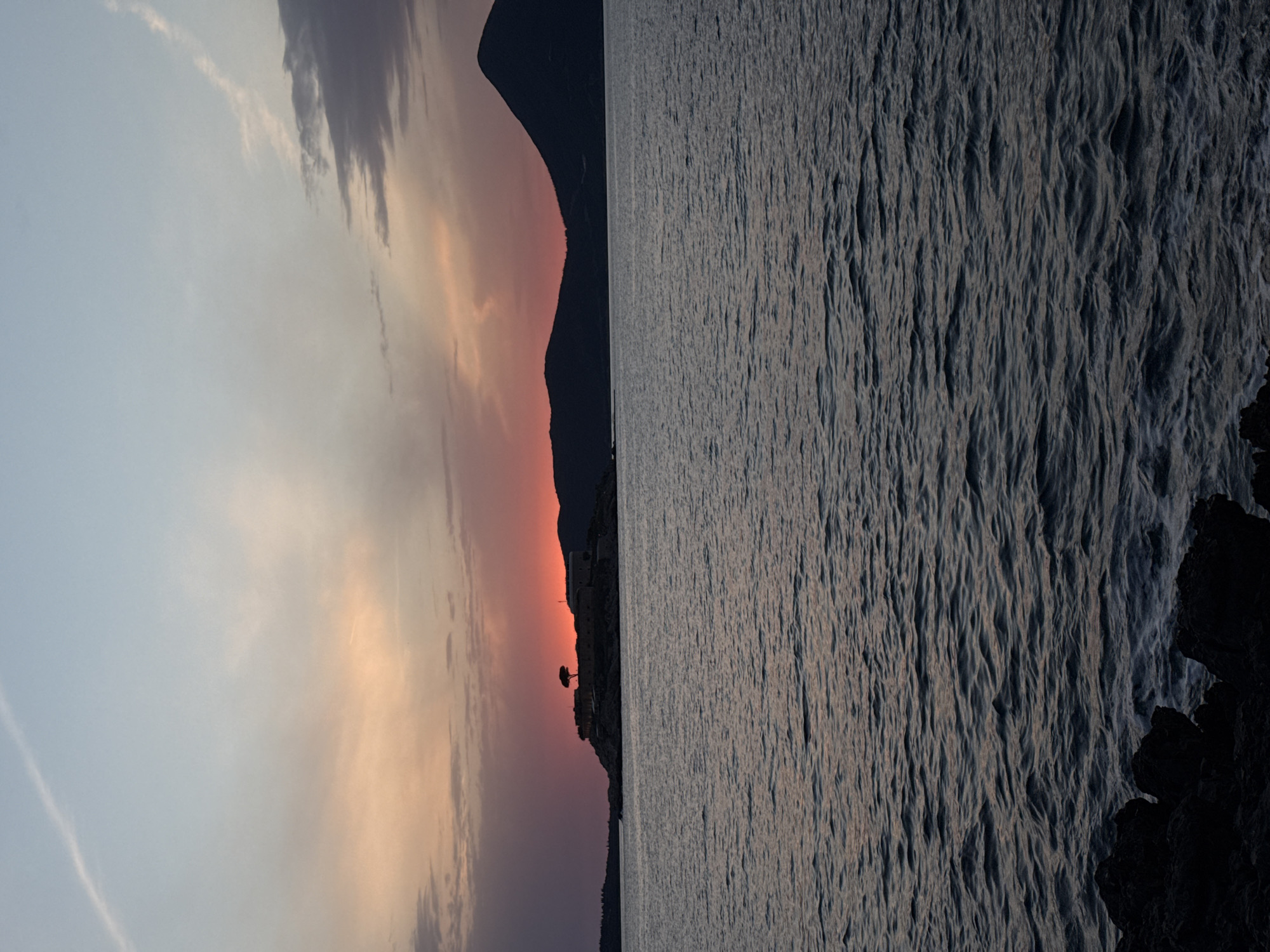 The image depicts a coastal landscape at sunset. The right side shows the sea with small waves, while the left side features the sky transitioning from light blue to orange near the horizon. Silhouetted mountains and a fortified structure with a lone tree are visible against the colorful sky. The image is likely taken near Herceg Novi, Montenegro, given the coastal landscape and architecture. A dark rocky cliff is in the lower right corner of the image.