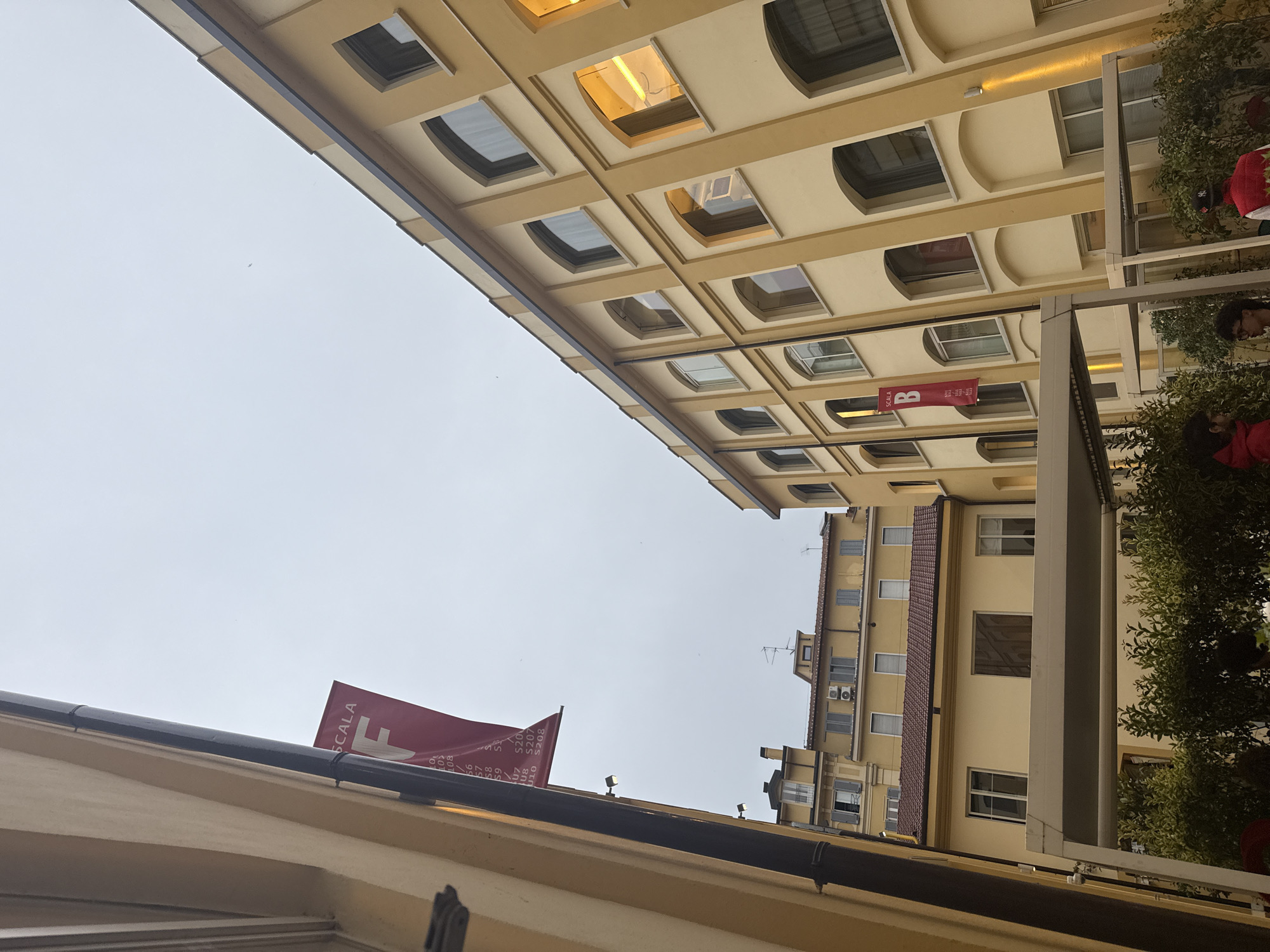 The image shows a view upwards between buildings in Milan, Italy. The sky is overcast. Two yellow buildings are visible, with regular window patterns on the left building. A banner for "SCALA" is visible in two locations: one red banner flying from the lower building, and a smaller banner between two buildings in the center of the image. Some green foliage is seen on the right side of the image. Part of a person is visible in the foliage.
