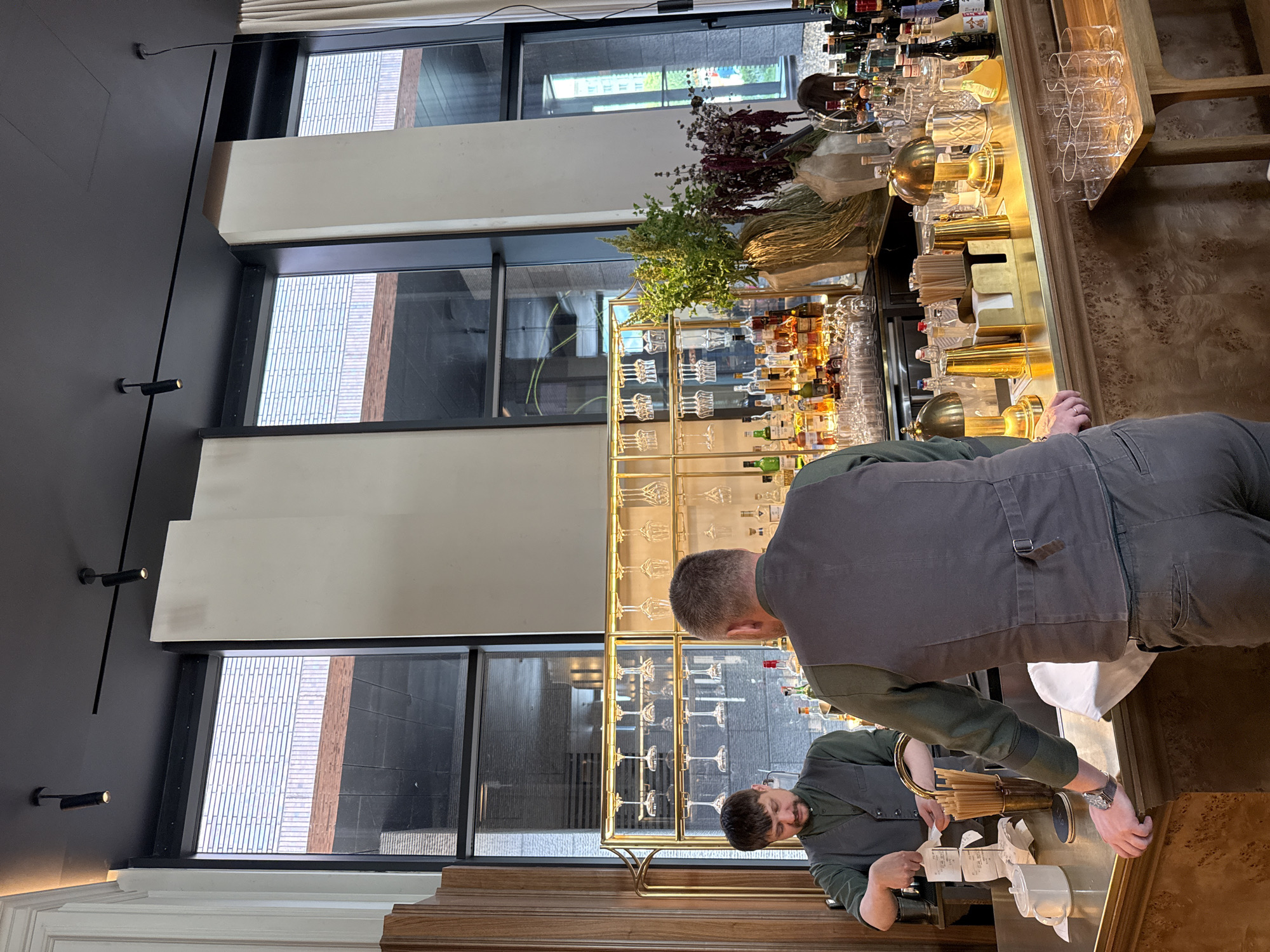 The image depicts the interior of a bar in Moscow, Russia. Two bartenders, dressed in dark vests over green shirts, are visible behind a polished wooden bar counter. One bartender on the left is handling receipts, while the other, facing away from the camera, leans on the counter. The bar area features a display of various bottles of liquor, wine glasses hanging from racks, and decorative vases with foliage. Large windows overlook an urban exterior with brick buildings. Spotlights are mounted on the dark ceiling, and a modern aesthetic is evident in the design elements.