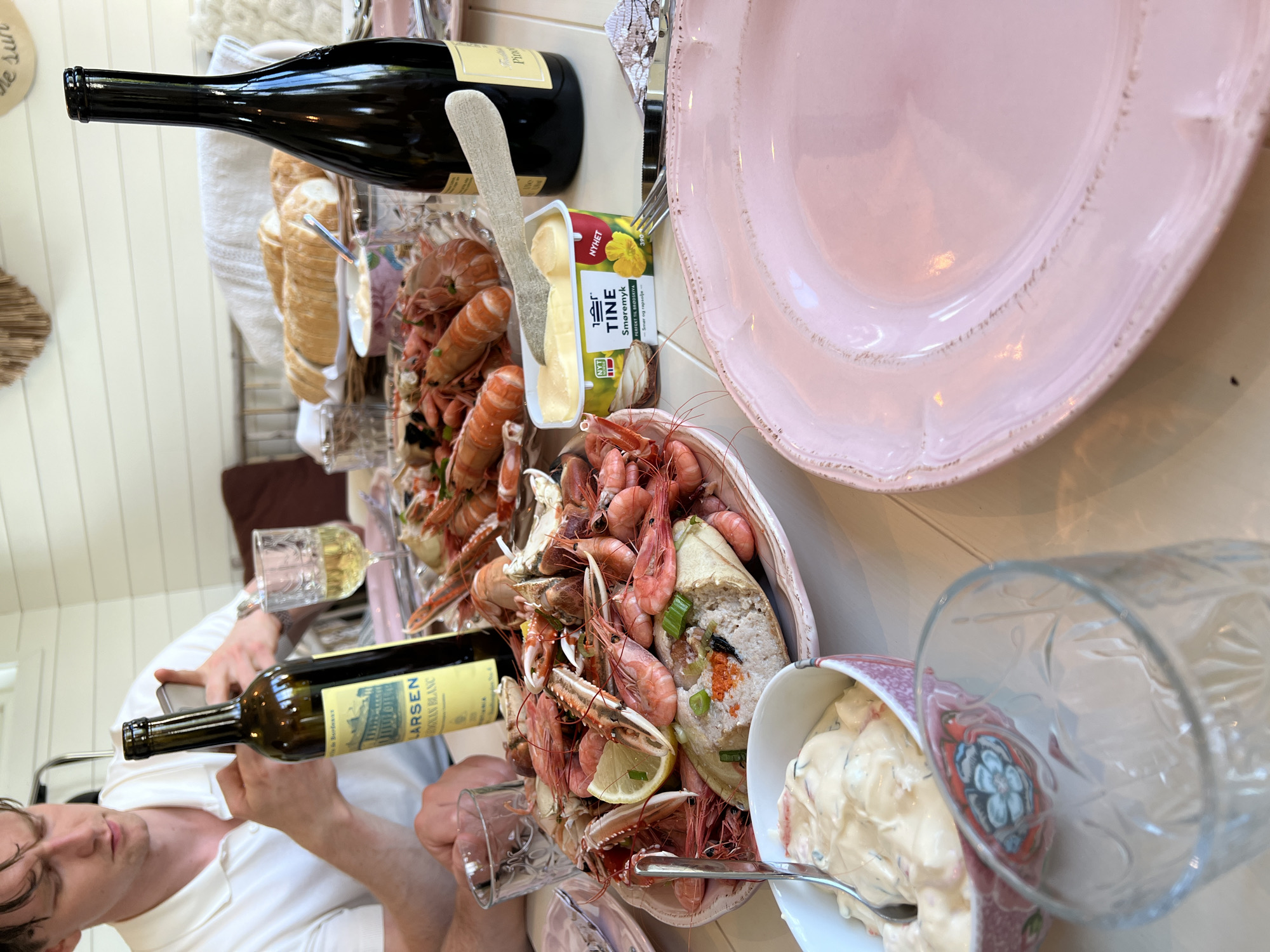 A person in a white polo shirt sits at a table, holding a phone. In front of them is a platter filled with cooked shrimp and langoustines, a sliced baguette, a small bowl of what appears to be mayonnaise-based dip, and a tub of butter. Two bottles of wine and several glasses are also on the table. The setting appears to be outdoors, possibly on a patio or deck. The image includes a hint of the geographical location in Larkollen, Norway, suggested by the context of the image.