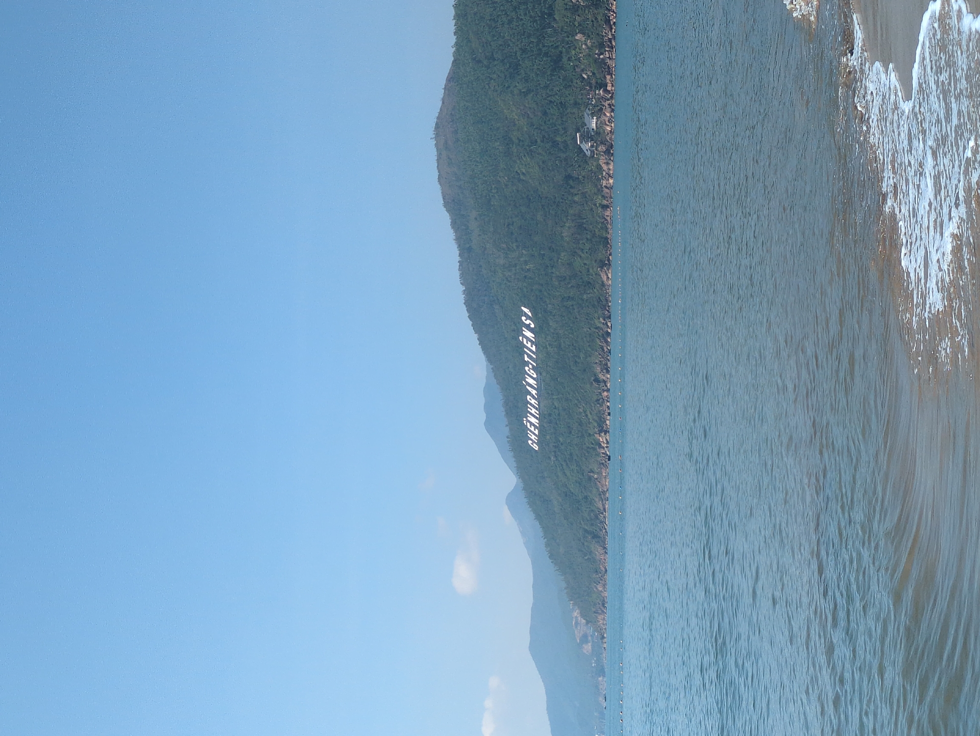 The image displays a coastal landscape within South Quy Nhon Ward, Vietnam. The scene is dominated by a clear, light blue sky containing one distant wispy cloud. A prominent range of lush green, forested mountains extends across the background, with large white capital letters spelling "GHENHRANG-TIEN SA" fixed to the slope of the central peak. In the midground, calm, blue-green ocean water features a horizontal line of white spherical buoys and several small, dark, unidentified objects. The foreground reveals a light brown sandy beach on the right, where small waves break into white foam. A multi-story building with a dark roof is visible nestled among the trees at the base of the mountain on the far right. No individuals are depicted in the scene.