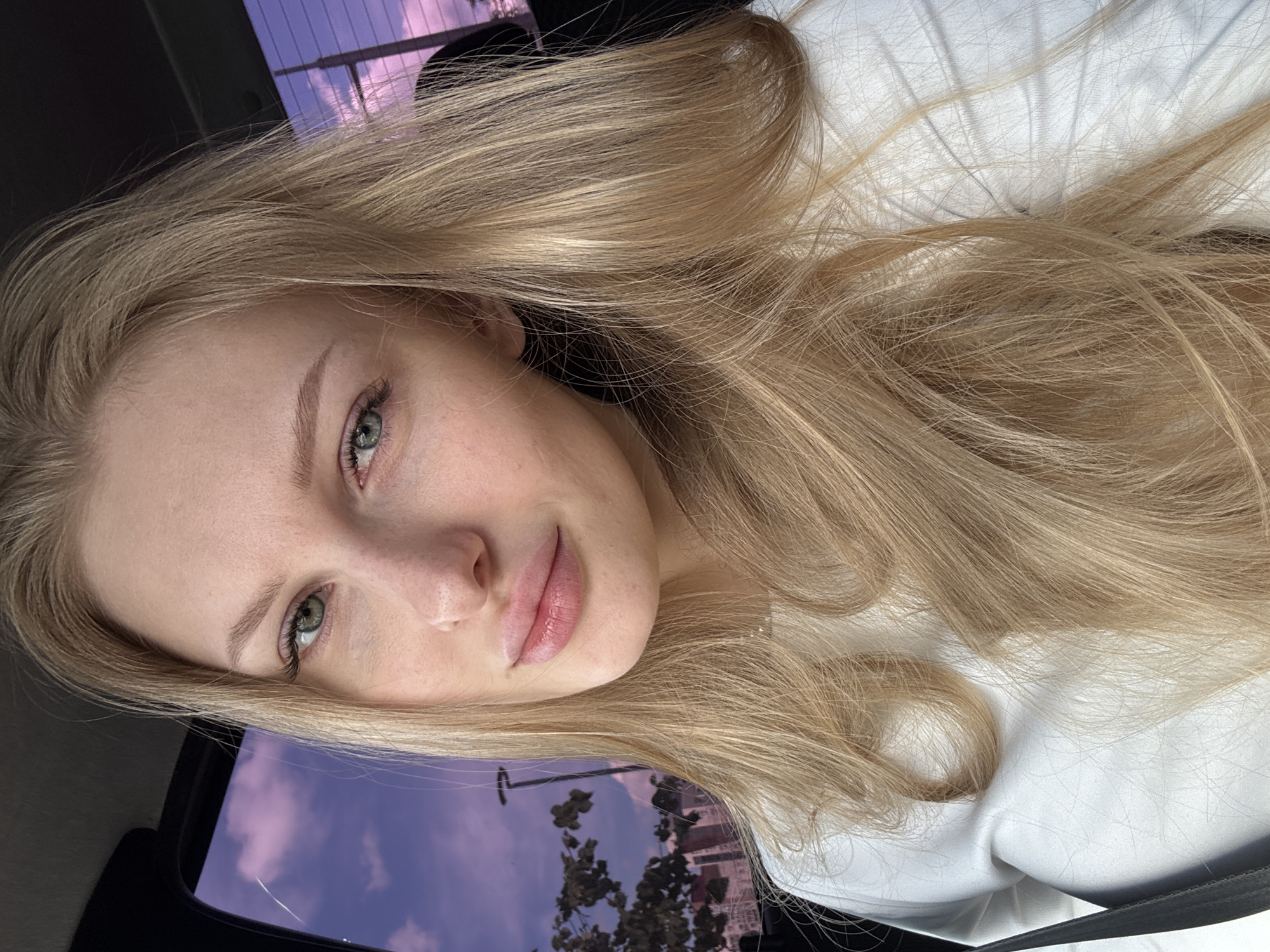 The image is a close-up selfie of a young woman with long, wavy blonde hair and clear light green eyes. Her head is tilted to the left, and she gazes towards the viewer with a soft, relaxed expression and a subtle, gentle smile. She has natural-looking makeup, highlighting her eyebrows, lashes, and lips with a delicate pink hue. She is wearing a light-colored, possibly white, top or jacket.
The woman appears to be inside a vehicle, with reflections and a view through a window visible in the background. Outside, a striking purple and pink sky with scattered clouds suggests either sunrise or sunset. Below the sky, indistinct silhouettes of trees and distant buildings are faintly visible, indicating an urban or suburban setting. The overall mood is serene and personal, capturing a quiet moment. No visible text is present in the image. The location is specified as Perm, Russia.