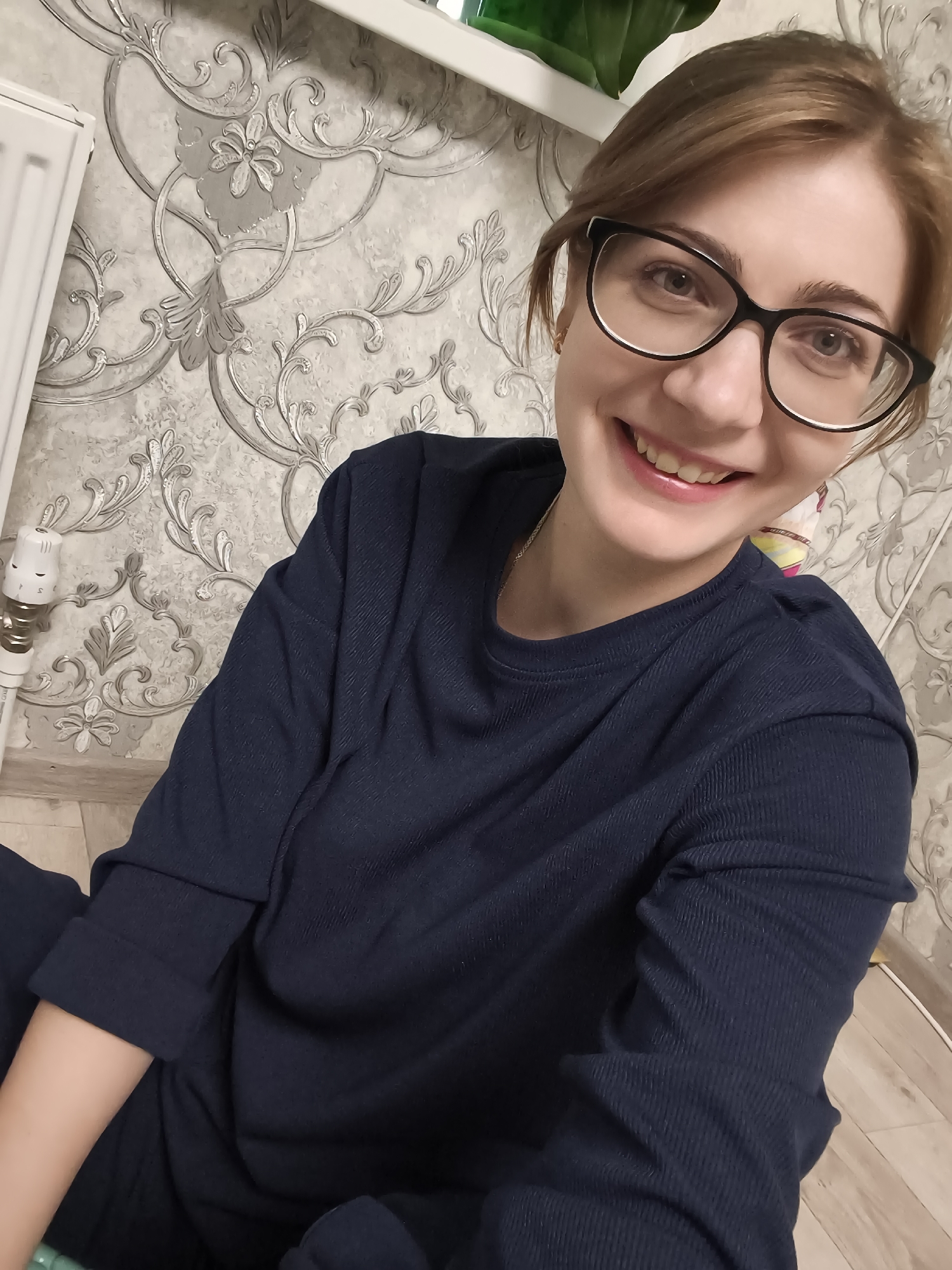 This is a selfie taken indoors, likely in a home in Podkletnoe, Russia. The subject is a woman with reddish-brown hair pulled back, wearing glasses with black frames and a dark navy blue long-sleeved shirt. She is smiling widely and looking directly at the camera.
The background features ornate wallpaper with a swirling, damask-like pattern in shades of grey and white. To the left of the woman, a white radiator is visible, attached to a wall with a thermostat. The floor appears to be made of light-colored wooden planks. In the upper right corner, a green leafy plant can be seen, suggesting the presence of houseplants.
The lighting suggests it is daytime, possibly late afternoon or evening, as the indoor lighting seems to be the primary light source. The overall mood conveyed is cheerful and relaxed. There is no visible text in the image.