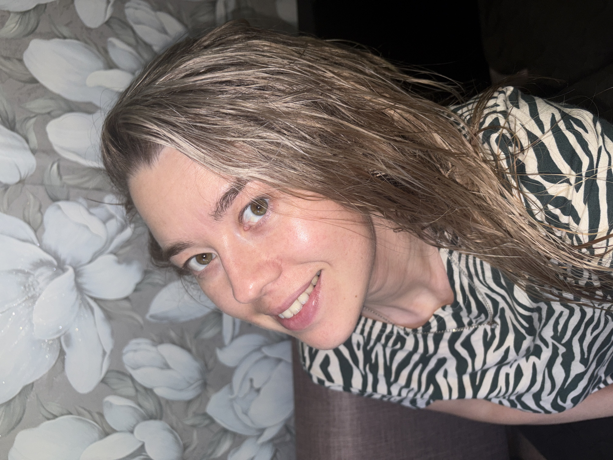 A woman is posing for a picture, looking up and smiling slightly. She has light brown hair that appears wet and is wearing a zebra-striped top. Behind her is a wall with a pattern of large white flowers on a grey background. The location is not discernible from the image provided, so it's impossible to confirm that it was taken in Chaikovsky, Russia.