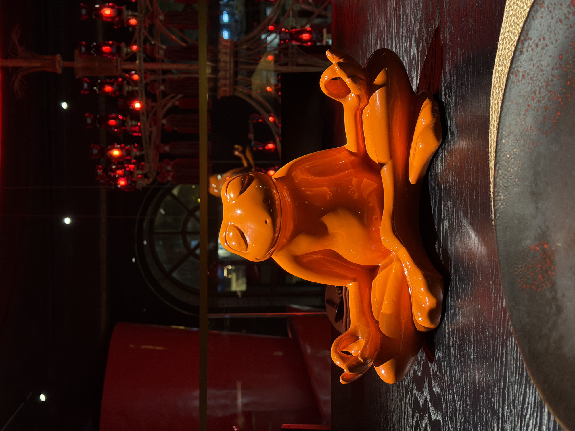 An eye-level indoor shot shows a glossy orange frog figure atop a dark, wood-grained surface. The frog has an anthropomorphic quality, with one hand gesturing in a circular shape above its head, while the other hand mirrors this gesture below its body.
In the blurred background, a chandelier-like structure with multiple red lights is visible, as well as a circular window with a grid pattern. A section of red furniture is also discernible.
The scene is potentially located in a restaurant or bar in Moscow, Russia, given the decor and atmosphere.