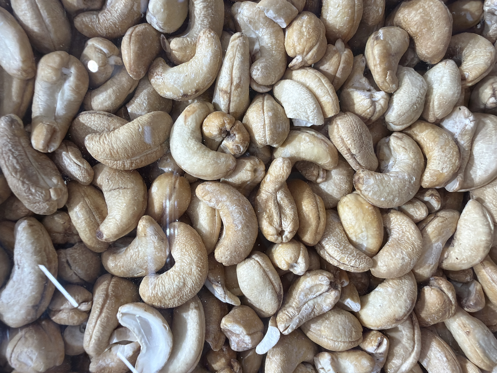 This is a close-up, overhead, eye-level shot of a large pile of roasted cashews. The cashews are a light to medium brown color with some white powdery residue on their surfaces, indicating they may be lightly salted or coated. They are irregularly shaped, with many exhibiting the characteristic kidney bean shape. Some cashews are whole, while others appear broken or partially shelled. The lighting suggests an indoor environment, possibly a market stall or food vendor. There are no people, animals, or specific landmarks visible in the image that would allow for identification of a location in Đà Nẵng, Vietnam. The focus is entirely on the texture and arrangement of the nuts.