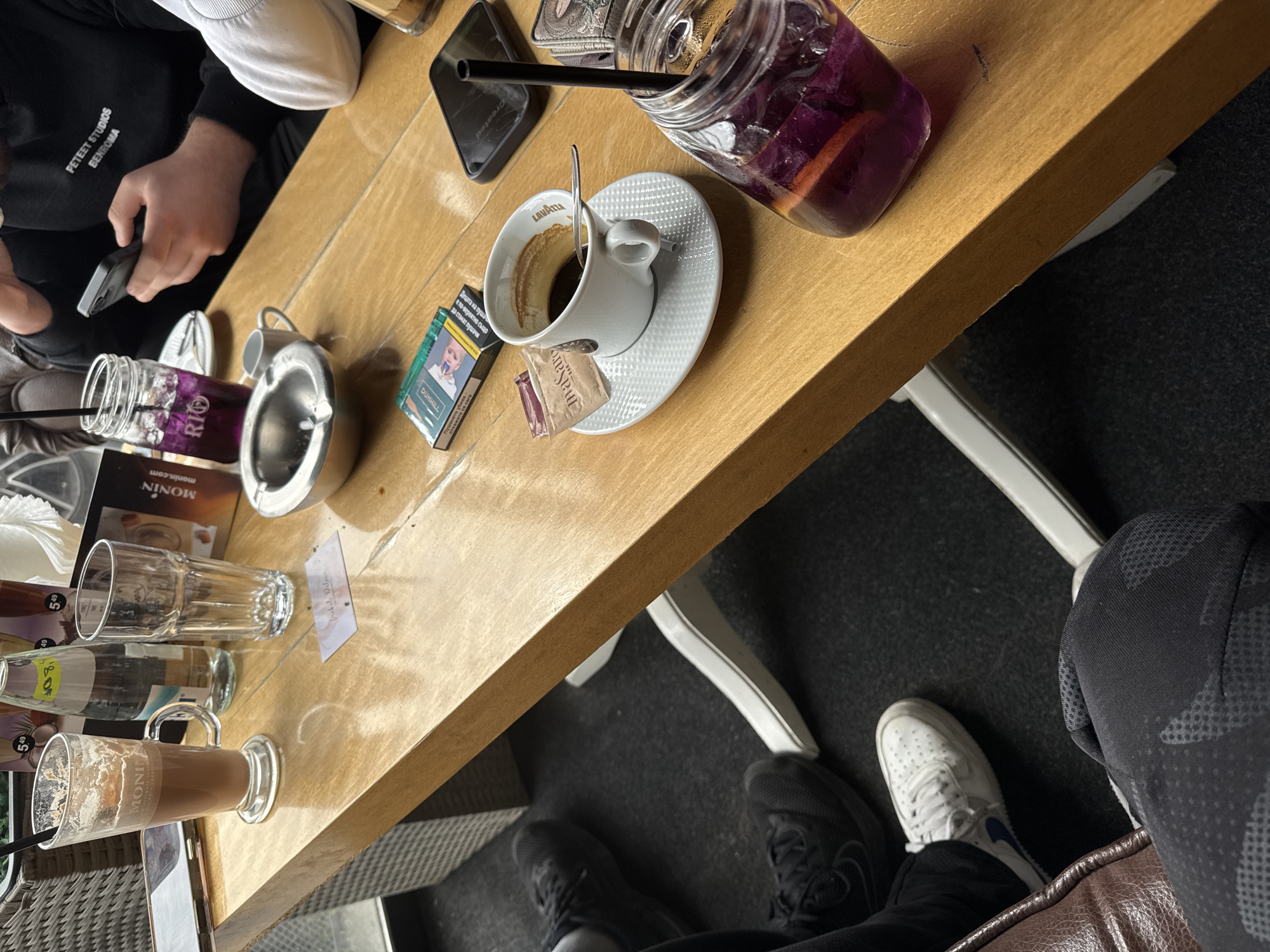 This image depicts a tabletop scene, likely at a cafe or restaurant, with several items commonly found in such a setting. The surface is a light-colored wooden table, around which at least two people are seated.
On the table, there is a partially consumed cup of Lavazza-branded coffee on a saucer with a spoon and sugar packets nearby. A pack of Dunhill cigarettes is also present. Additionally, there are multiple glass beverages including a jar drink with a straw, possibly a violet-colored cocktail, and other glasses, one appearing to contain a Monin-branded drink. An ashtray is also visible.
Parts of at least two people are visible at the table: one person holding a mobile phone and wearing a cap and another person's legs and feet, including one foot with a white sneaker and another with a black sneaker.
The scene appears to be casual. Without further information, determining if it is a café in Varna, Bulgaria, is not possible.