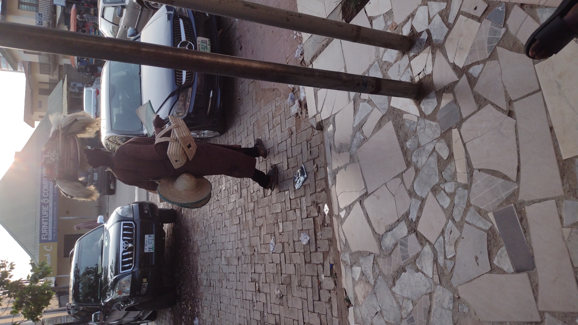 A man in traditional Nigerian attire, including a feathered headpiece, is walking down a street in Jos, Nigeria. He carries woven goods. Several cars are parked along the side of the road. One black SUV has a license plate that reads "KWL-323JM." Another car has a license plate that reads "ABUJA 92JD." A building is visible in the background with a sign that says "Furniture & Computers." The street is paved with a mix of brick and tile patterns. The image is rotated clockwise.