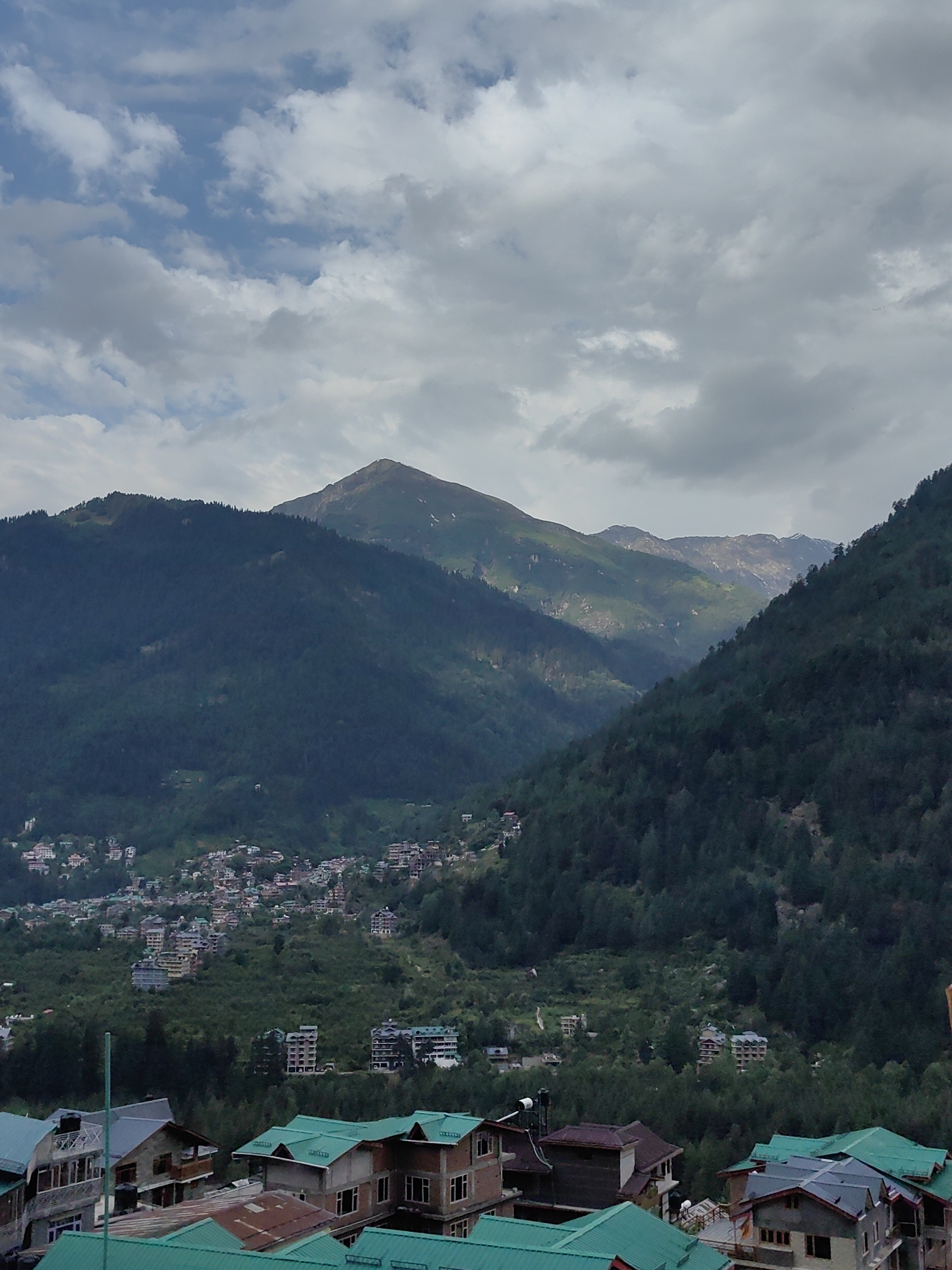 The image captures a scenic view of a mountainous region with a town nestled within the valley. In the foreground, several buildings with distinctive green, sloped roofs are visible, suggesting a residential or commercial area. These buildings have multiple stories and appear to be constructed from brick and concrete.
Beyond the foreground buildings, the landscape transitions to a densely forested mountainside. The trees are predominantly dark green, indicating a lush, temperate or subtropical environment. Scattered throughout the hillside are clusters of buildings, forming a town or village that extends up the slopes. These buildings are varied in size and architectural style, with some appearing to be multi-story apartment complexes and others smaller houses.
Further in the background, larger mountain ranges dominate the scene. One prominent peak in the center-right of the image is topped with snow, even though the lower slopes are covered in vegetation. The sky above is partly cloudy, with patches of blue visible between masses of grey and white clouds. The overall impression is of a tranquil, elevated settlement surrounded by natural grandeur. Given the architectural style of the buildings, particularly the green roofs, and the mountainous terrain, the location strongly resembles the town of Vashisht, India, which is known for its hot springs and its proximity to the Himalayas.