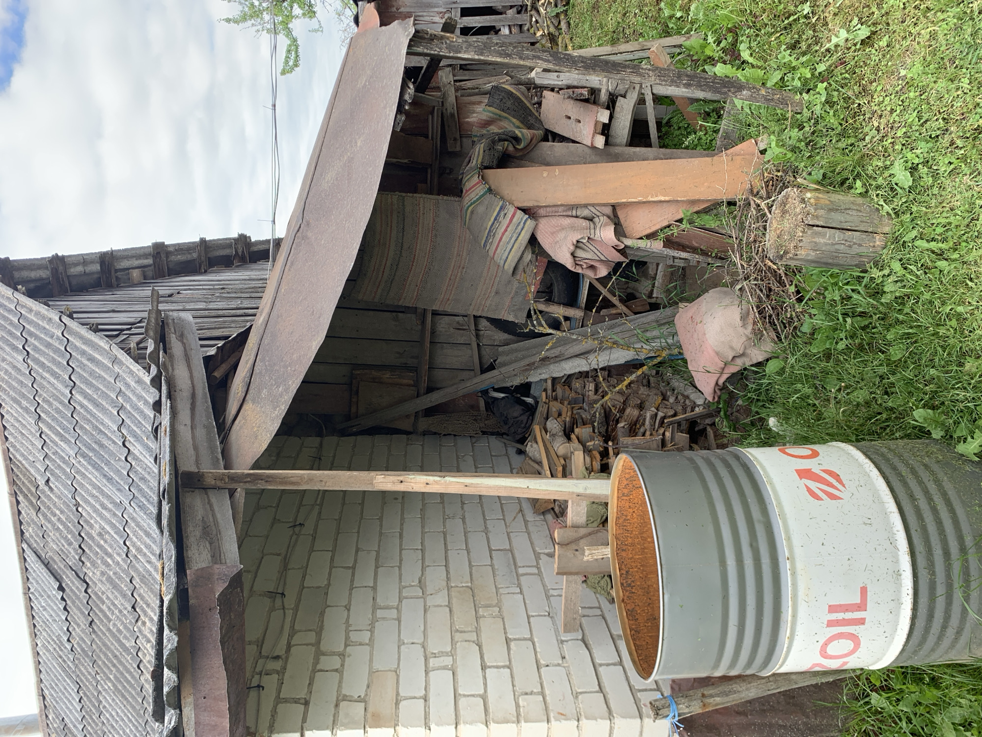 This image captures a weathered rural utility area in Відзаўскі сельскі Савет, Belarus, under an overcast sky. On the left, a structure with a white brick facade features a grey, corrugated asbestos-cement roof exhibiting signs of age and minor discoloration. Beneath the main roof, a rusted sheet metal gutter or flashing is affixed to a wooden support beam.
Adjacent to the brick structure, a lean-to or shed constructed from aged, dark wooden planks stands on the right. This lean-to is covered by a sloped roof of heavily rusted corrugated metal. Underneath this shelter, various items are stored, including a stack of cut firewood and timber, and several draped or folded fabric items, notably two patterned, striped rugs or blankets. Additional wooden boards, debris, and a partially obscured dark bag are also present within the lean-to.
In the foreground, a large, open-top metal barrel is positioned on the left. Its lower section is grey, while a central white band displays the word "OIL" in red capital letters, accompanied by a red stylized emblem. The interior of the barrel shows significant orange-brown rust. The ground is covered with green grass and assorted weeds. A weathered wooden tree stump is centrally located in the lower portion of the frame, surrounded by loose twigs and branches. In the upper right background, two horizontally strung electrical wires are visible against the cloudy sky, alongside a small portion of green tree foliage. No individuals or animate subjects are present.