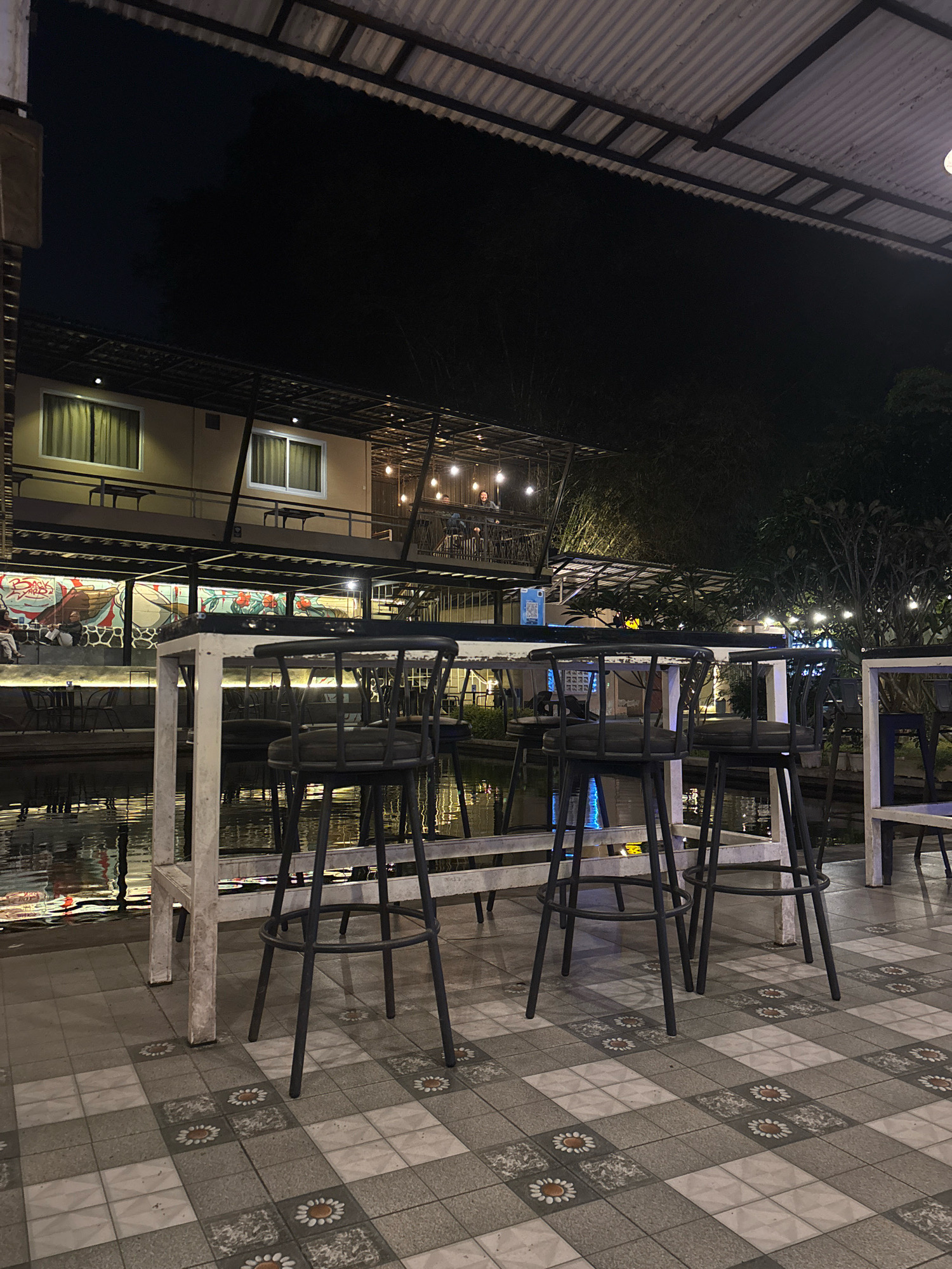 This is an exterior night shot of a restaurant with a water feature. The restaurant is a two-story building with a corrugated metal roof overhang. The first floor has a mural and several tables and chairs visible through a large open area that overlooks the water. The second floor has windows with curtains and a balcony with people. In the foreground, there are several bar stools and tables placed on a tiled patio. The water feature is reflecting the lights from the restaurant and string lights are visible throughout the scene. The scene appears to be in Sukabumi, Indonesia.
