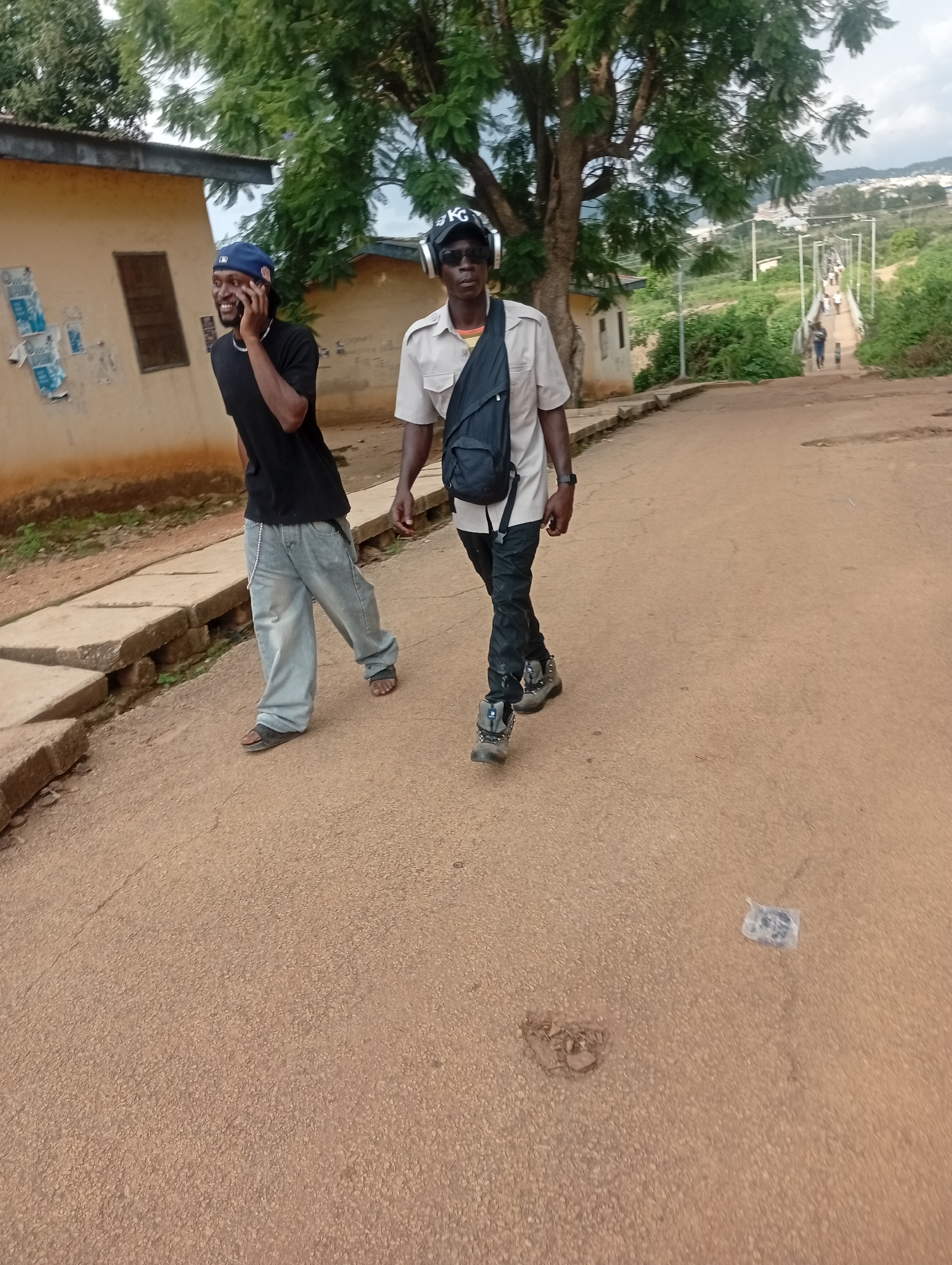 Two men are walking on a paved road in Rafin Par, Nigeria. The man on the left is wearing a black t-shirt, light blue baggy jeans, and sandals, and is talking on a cell phone. The man on the right is wearing a light-colored short-sleeved shirt, dark pants, and hiking boots, and has headphones on. He is also wearing a backpack. The road is lined with concrete barriers and vegetation. In the background, there are buildings and a bridge with people walking on it. The sky is overcast.