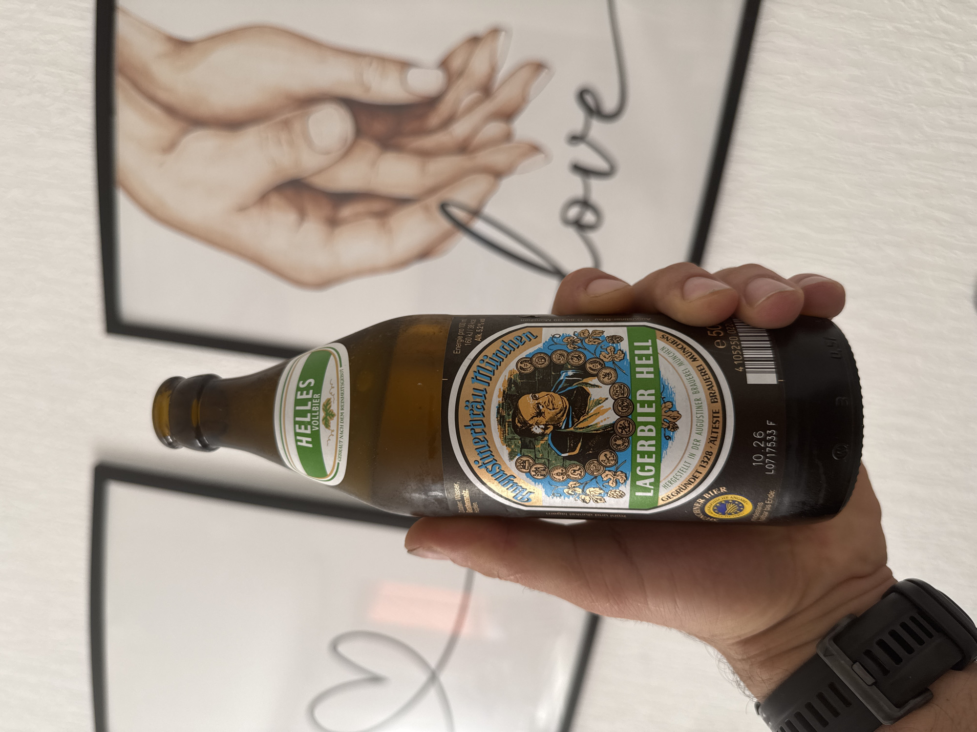 A person is holding a brown glass beer bottle in their hand, with a black sports watch visible on their wrist. The beer is Augustinerbräu München Lagerbier Hell. The label states the beer is brewed in Munich according to the Reinheitsgebot (purity law) and contains water, barley malt, and hops. The bottle's expiration date is listed as 10.26, with the code L0717533 F. The beer is a Vollbier Helles. The background includes a framed picture featuring a drawing of human hands forming a heart shape, with the word "love" written above. A second framed picture is behind. The scene appears to be indoors. The city of Füssen, Germany, is not mentioned or depicted in the image.