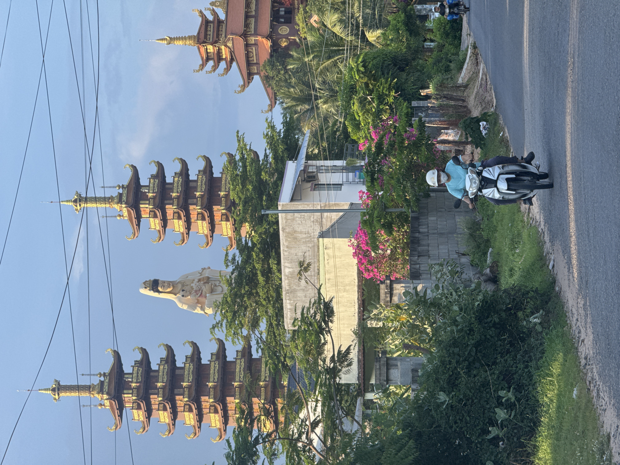 The image depicts a roadside scene in Mui Ne, Vietnam. In the background, several towering, multi-tiered pagodas with golden roofs are visible, along with a large white statue of a seated figure holding a child. Lush green trees and vegetation surround the structures. In the foreground, a person wearing a light blue shirt, dark pants, and a white helmet is riding a white motorcycle down an asphalt road. They are wearing a light blue face mask. Further down the road, another person on a motorcycle is visible in the distance. The environment includes a grey concrete wall, various plants, and an overcast sky.