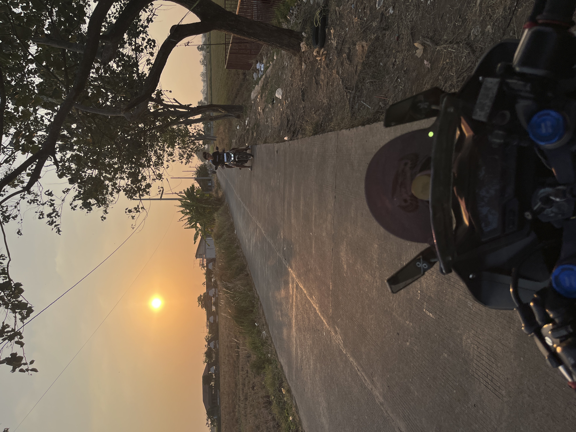 A first-person perspective shot shows a person on a motorcycle driving on a paved road in the rural outskirts of Pecangaan, Indonesia, during sunset. The sun is low in the sky, casting a warm, orange glow over the scene. To the left of the road are harvested rice fields and some buildings with corrugated roofs. To the right of the road are trees, dirt, and a rusty fence. A single person is riding a motorcycle further down the road, and the camera's motorcycle also has a person on it. The sky is a gradient of orange and yellow.
