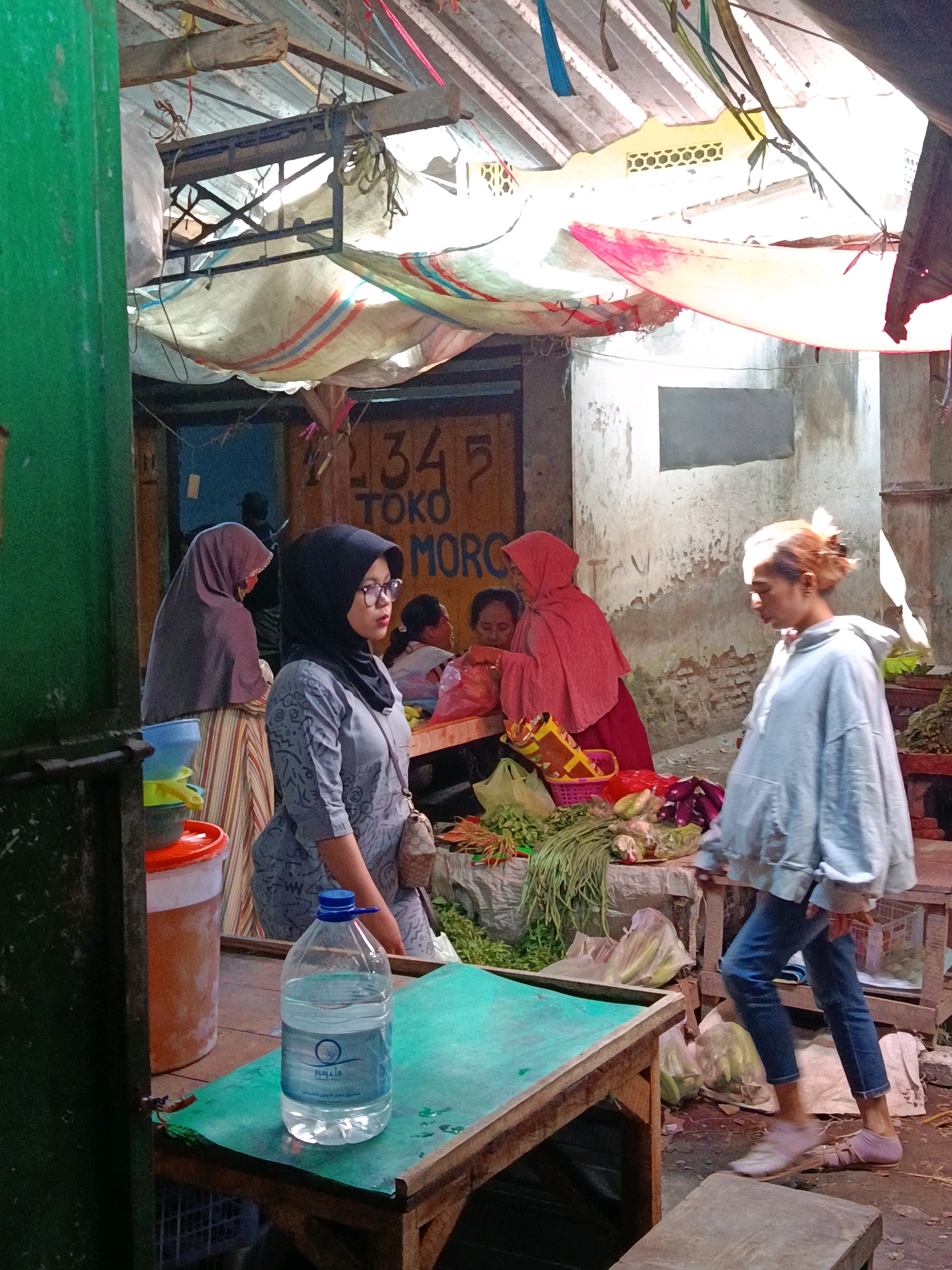 This image depicts a busy outdoor market stall in Jatiroto, Indonesia. The scene is crowded with people and goods, suggesting a vibrant commercial atmosphere.
In the foreground, a woman wearing a black hijab and glasses stands by a table. She is dressed in a patterned grey tunic and has a small patterned bag slung over her shoulder. To her left, a large plastic bottle of water with a blue cap sits on a green surface, which appears to be part of a wooden table. Behind her, another woman wearing a pink hijab is visible, along with other shoppers and market vendors.
The market stall itself is covered by a makeshift roof made of tarps and fabric, creating a shaded area. Various fresh produce, including vegetables like long beans and what appear to be eggplants, are displayed on tables and in baskets. The background reveals more stalls and structures, with some signage that includes the numbers "34" and the word "TOKO" (shop in Indonesian). The overall environment suggests a typical Indonesian market, with a mix of goods, people, and rudimentary shelter.