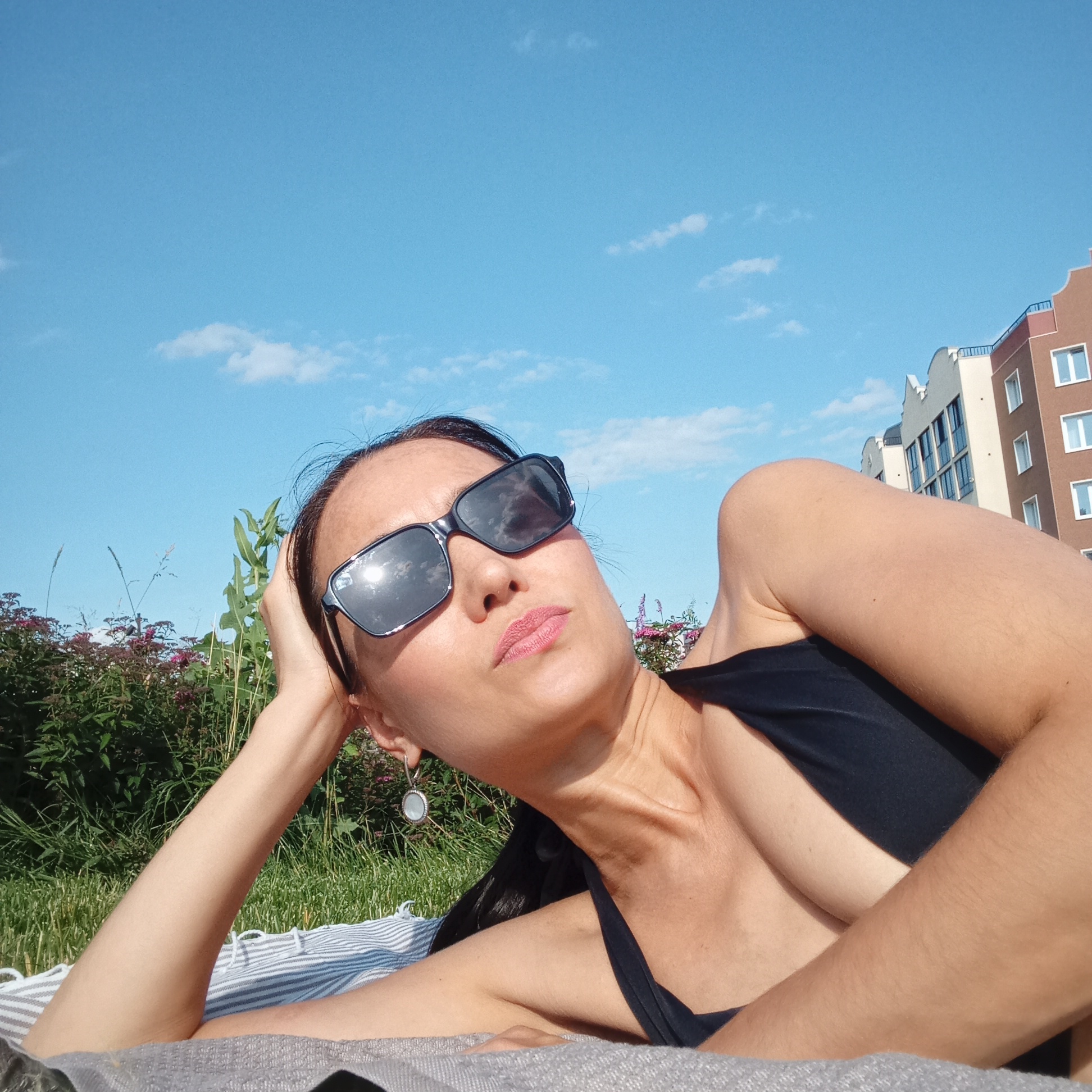 A woman lies on a towel outdoors, wearing sunglasses and a black swimsuit. She is resting her head on her arm, with her body angled away from the camera. In the background, there is green grass, bushes with pink flowers, a blue sky with scattered clouds, and a multi-story building with cream and brown facades. The scene is lit by daylight, casting shadows. The woman appears to be in a relaxed pose, possibly sunbathing. The location is identified as the city of Pervomaysky, Russia.
