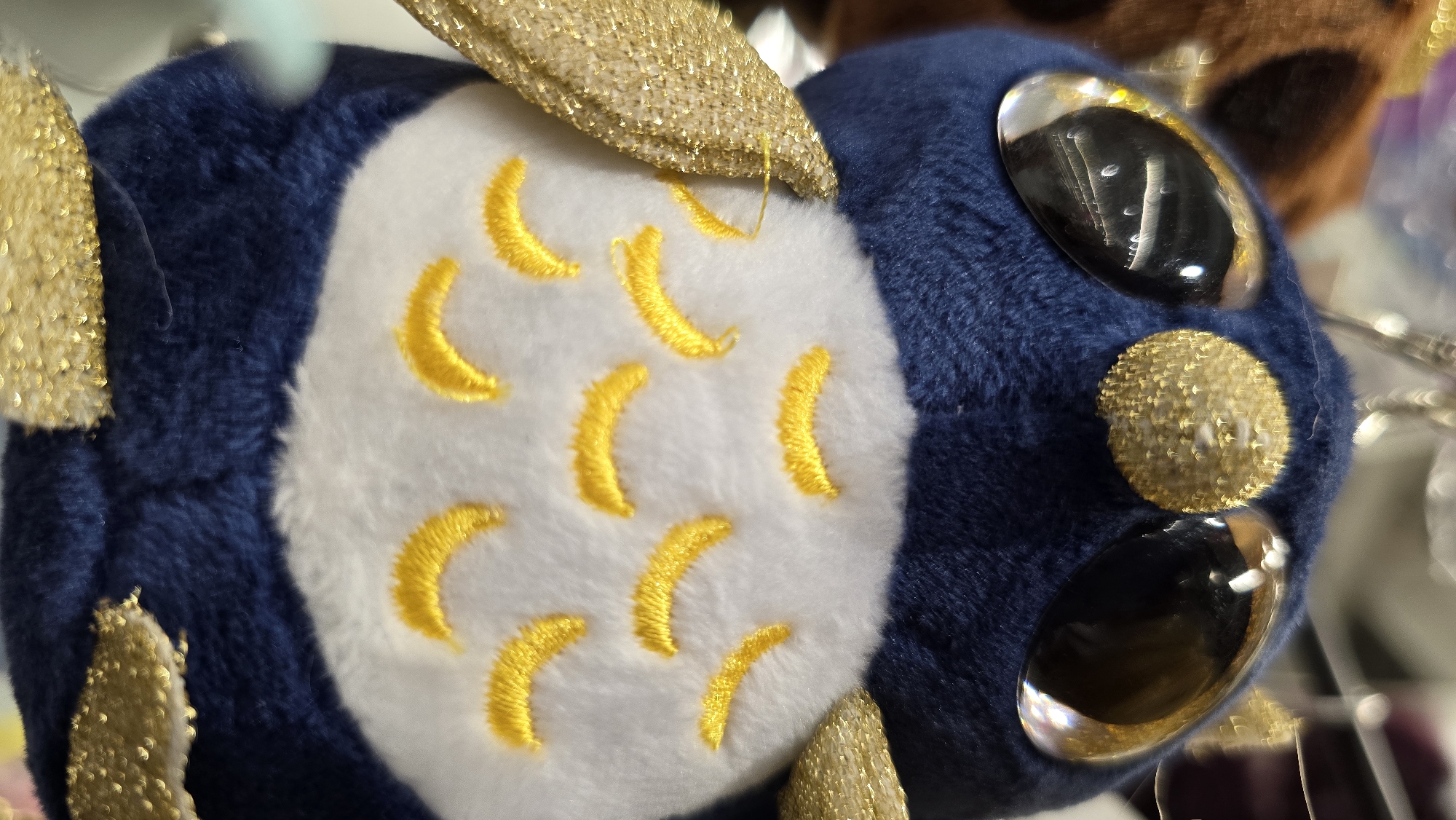 This is a close-up, high-angle, eye-level shot of a dark blue plush owl toy. The owl has large, reflective, black eyes with a golden sheen and a textured, golden, circular detail between its eyes. Its chest is white and embroidered with yellow crescent shapes, resembling feathers or scales. The owl has gold, glittery wings. The background is blurred and appears to be an indoor setting with some lights creating bokeh effects. The image does not depict any specific location in Krasnodar, Russia.