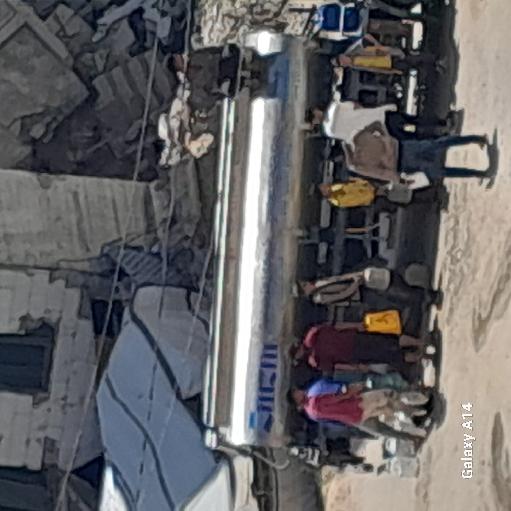 The image captures an outdoor scene, likely in Khan Yunis, Palestinian Territories, showing several individuals gathered around a large, horizontal, metallic cylindrical object. This object, possibly a water tanker or a large pipe, dominates the central part of the frame and features indistinct blue markings that could be text or a logo.
Multiple people, appearing to be adults and possibly children, are positioned on the right side of the cylindrical object, standing or sitting on the light-colored, sandy ground. They seem to be involved in an activity related to water collection or distribution, though specific containers are not clearly visible due to the image resolution. Some individuals are dressed in bright clothing, including hues of red, blue, and yellow.
The background on the left side of the image reveals structures or buildings that appear to be damaged or in disrepair, surrounded by rubble and debris, suggesting a challenging or conflict-affected environment. The ground throughout the scene is dusty and light brown. The lighting indicates it is daytime, with no strong shadows to suggest a specific time of day beyond broad daylight. The overall mood appears task-oriented, without clear expressions of emotion visible. In the lower right corner, the text "Galaxy A14" is visible, indicating the device used to capture the photograph.