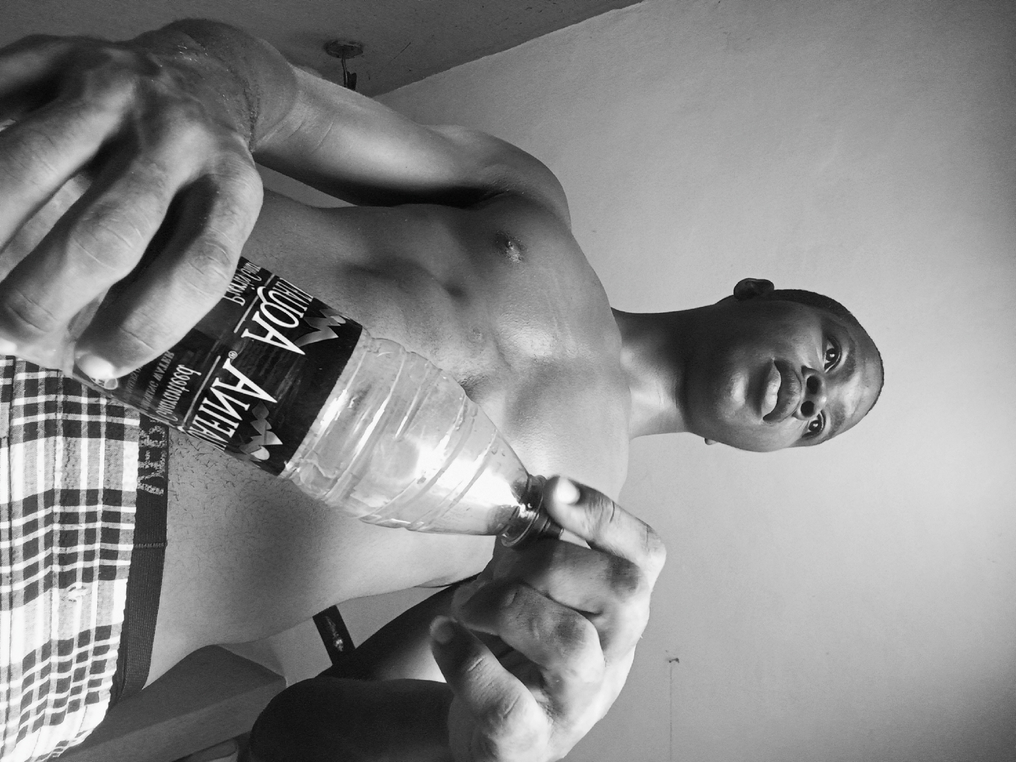 A black and white, low-angle shot depicts a muscular, shirtless man holding a plastic water bottle. He is wearing patterned shorts. The brand label on the water bottle is partially visible. The ceiling is visible in the background, and a light fixture is visible. The location is inferred to be Nkpor, Nigeria based on the prompt, although this is not visibly verifiable in the image itself.