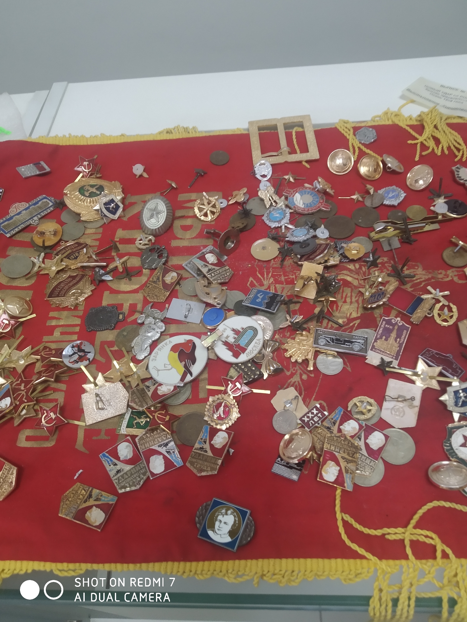 The image shows a collection of Soviet-era pins and medals spread out on a red cloth with a yellow fringe. The pins and medals feature various symbols, including stars, hammers and sickles, portraits of Lenin, and various organizational logos. Some of the pins also include inscriptions in Russian, such as "Udarnik" and "XXX Let."
There are a couple of pins that can be seen more clearly; one is from 1978 and features a communist youth organization logo and another is from a church in Kondopoga. Another features a bird called "Zaryanka".
The text at the bottom of the image indicates that the photo was taken with a Redmi 7 phone using AI Dual Camera.