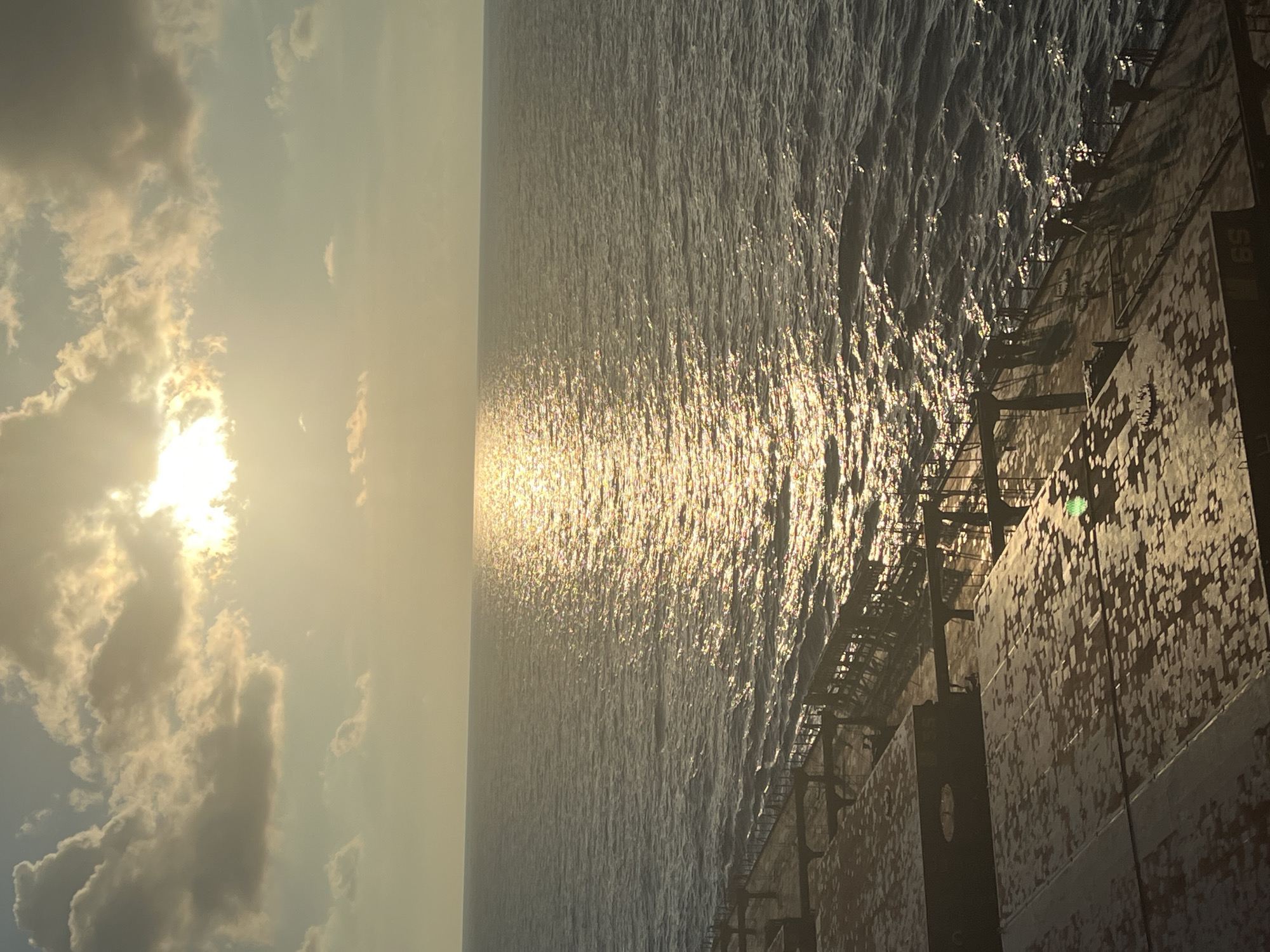 The image shows a view from a ship's deck looking out at the ocean during sunset. The sun is partially obscured by clouds, casting a golden and shimmering reflection on the water. The deck of the ship, with its metallic surfaces and weathered appearance, occupies the foreground. The ocean stretches out to the horizon, meeting a sky filled with dramatic clouds.