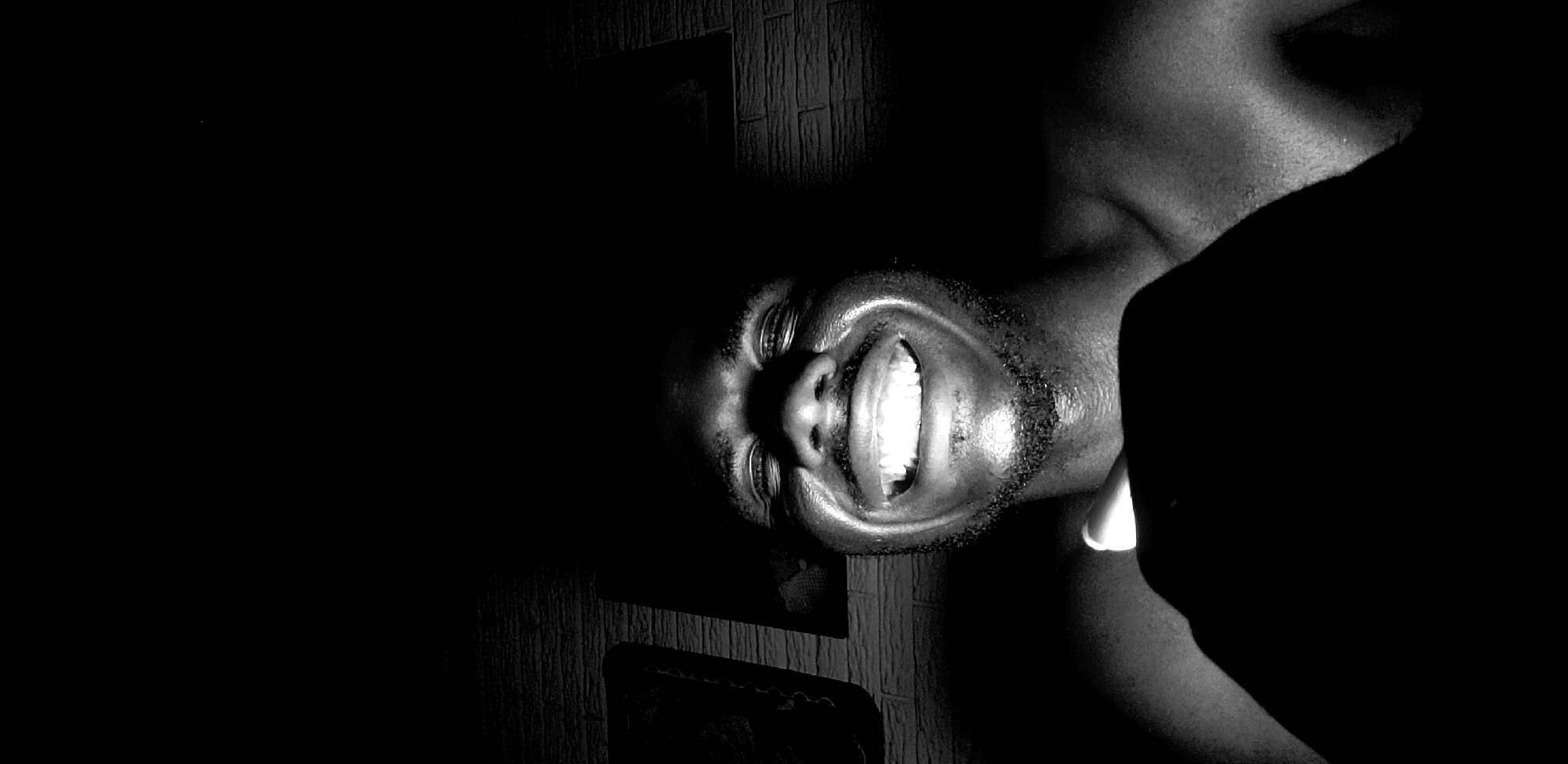 This monochromatic, close-up portrait features a person, likely male, depicted with their head tilted, appearing rotated. The individual's face and upper torso are illuminated by a stark, directional light source that casts dramatic highlights and deep shadows. The background is predominantly dark, with a textured wall, possibly wood paneling or brickwork, faintly visible in the upper left corner.
The person is captured in a moment of broad and joyful smiling, revealing a full set of prominent, white teeth. Their eyes are partially closed in the expression, and a sheen on the skin, possibly from sweat, reflects the intense light. Facial hair is visible around the chin and mouth area. The strong contrast between the illuminated subject and the surrounding darkness creates a dramatic and intimate atmosphere. No text is visible within the image. The scene appears to be an indoor setting, likely in a dimly lit environment, possibly at night, in Narayi, Nigeria.