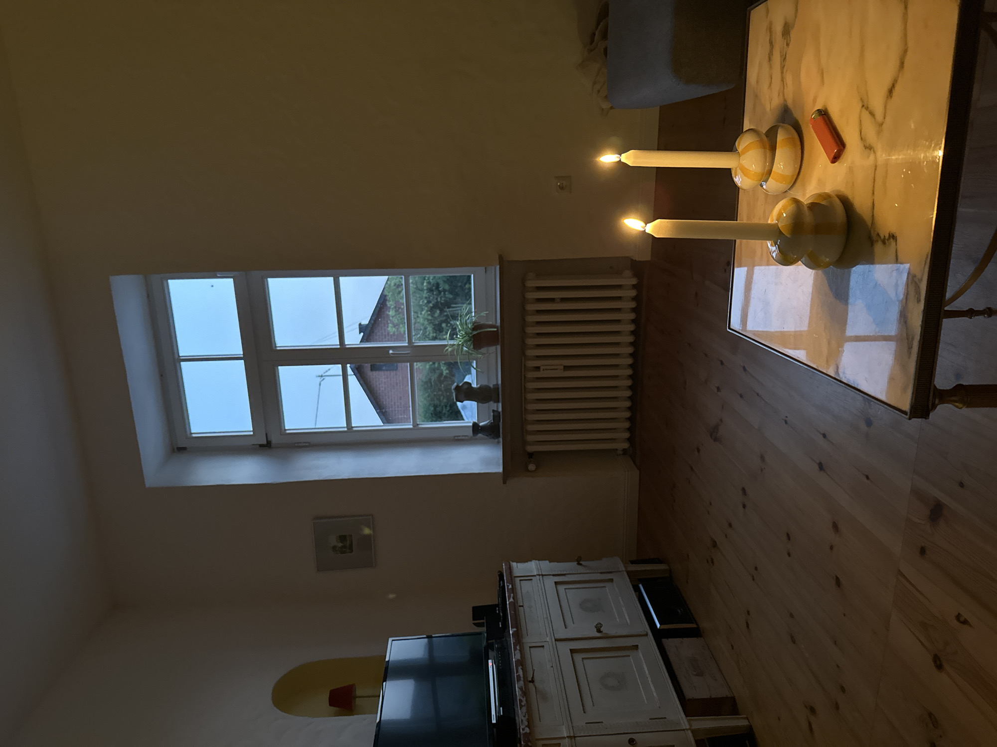 The image depicts a dimly lit interior scene, likely a living room, with a large window in the background. Outside the window, a brick house and trees are visible against a twilight sky. Inside, a white radiator is positioned beneath the window. Two lit candles in yellow and white patterned holders stand on a marble-topped coffee table, next to a red lighter. To the left of the window is a white dresser with closed cabinets, and a black television set is placed on top of it. A framed picture hangs on the wall above the dresser. The floor is made of wooden planks. The scene is consistent with a residence in Ammeldingen bei Neuerburg, Germany, given the architectural style and typical residential setting.