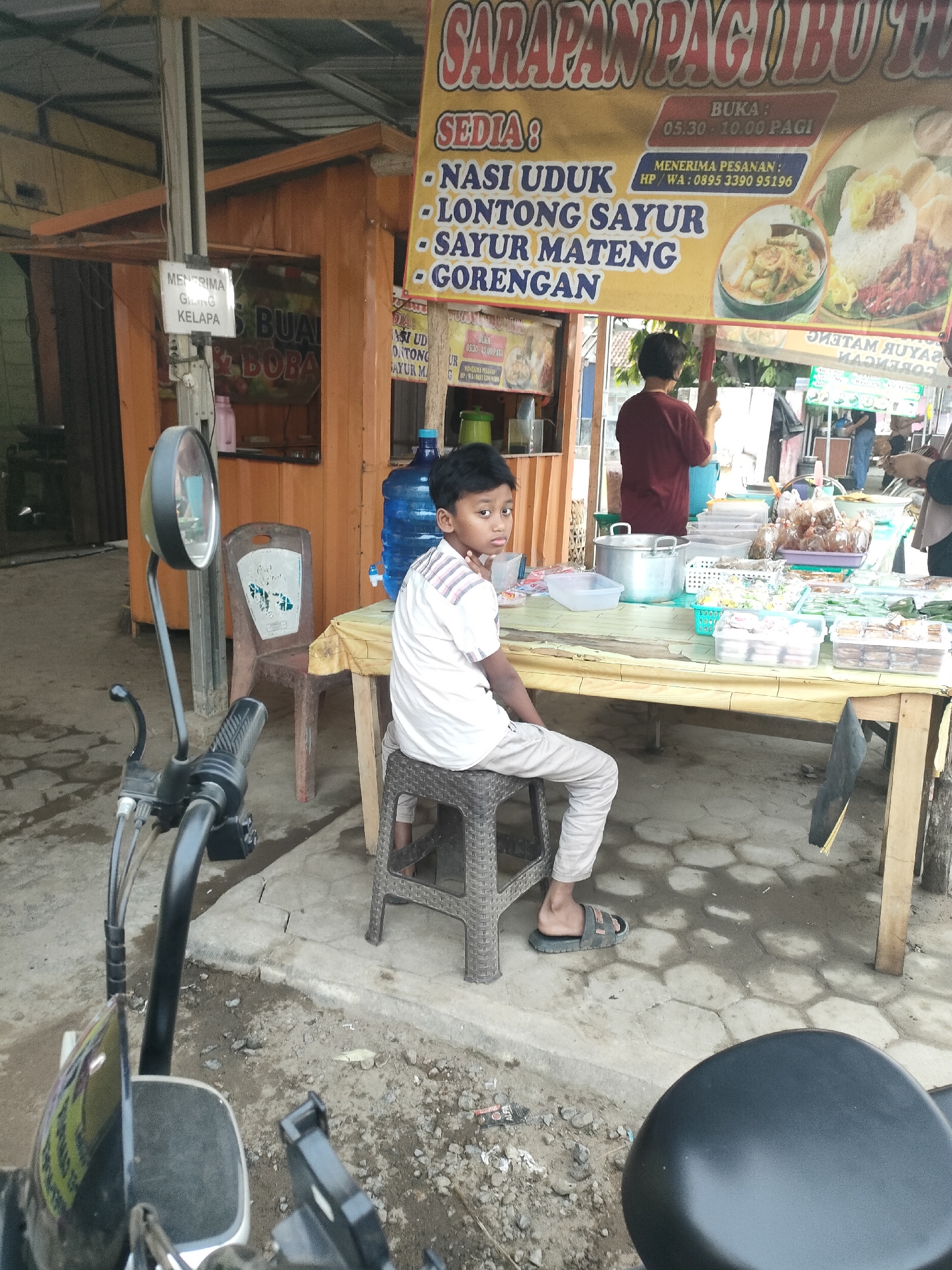 The image depicts an outdoor food stall with a young boy sitting on a stool. The stall is characterized by a yellow banner displaying food items and business hours. Various food items are laid out on a table, along with containers and a large water dispenser. A motorcycle is partially visible in the foreground. The background shows a wooden structure and other people in the distance. The setting appears to be a market or roadside eatery. The text on the banner, in Indonesian, indicates "SARAPAN PAGI ITU TU" (Breakfast is that) and lists menu items such as "NASI UDUK," "LONGTONG SAYUR," "SAYUR MATENG," and "GORENGAN." The business hours are listed as "BUKA: 05.30 - 10.00 PAGI" (Open: 05:30 - 10:00 AM). A phone number for orders is also visible. The presence of Indonesian text and the style of the food stall suggest this is located in Indonesia. The query specifies "Sukoyoso, Indonesia" as the location.