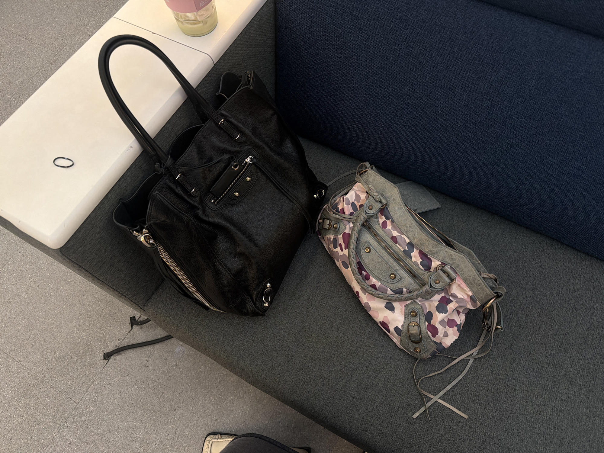 The image shows two handbags, one black and one gray patterned, resting on a gray upholstered surface next to a white side table with a transparent container on it. The black handbag is positioned closer to the side table, while the gray bag sits on the upholstery near a blue fabric surface. The flooring near the seating area is concrete or tiled. The bottom of a person wearing white-soled shoes is visible near the bottom of the frame. There is a black power cord visible. The exact location in Seoul, South Korea cannot be determined from this image alone.