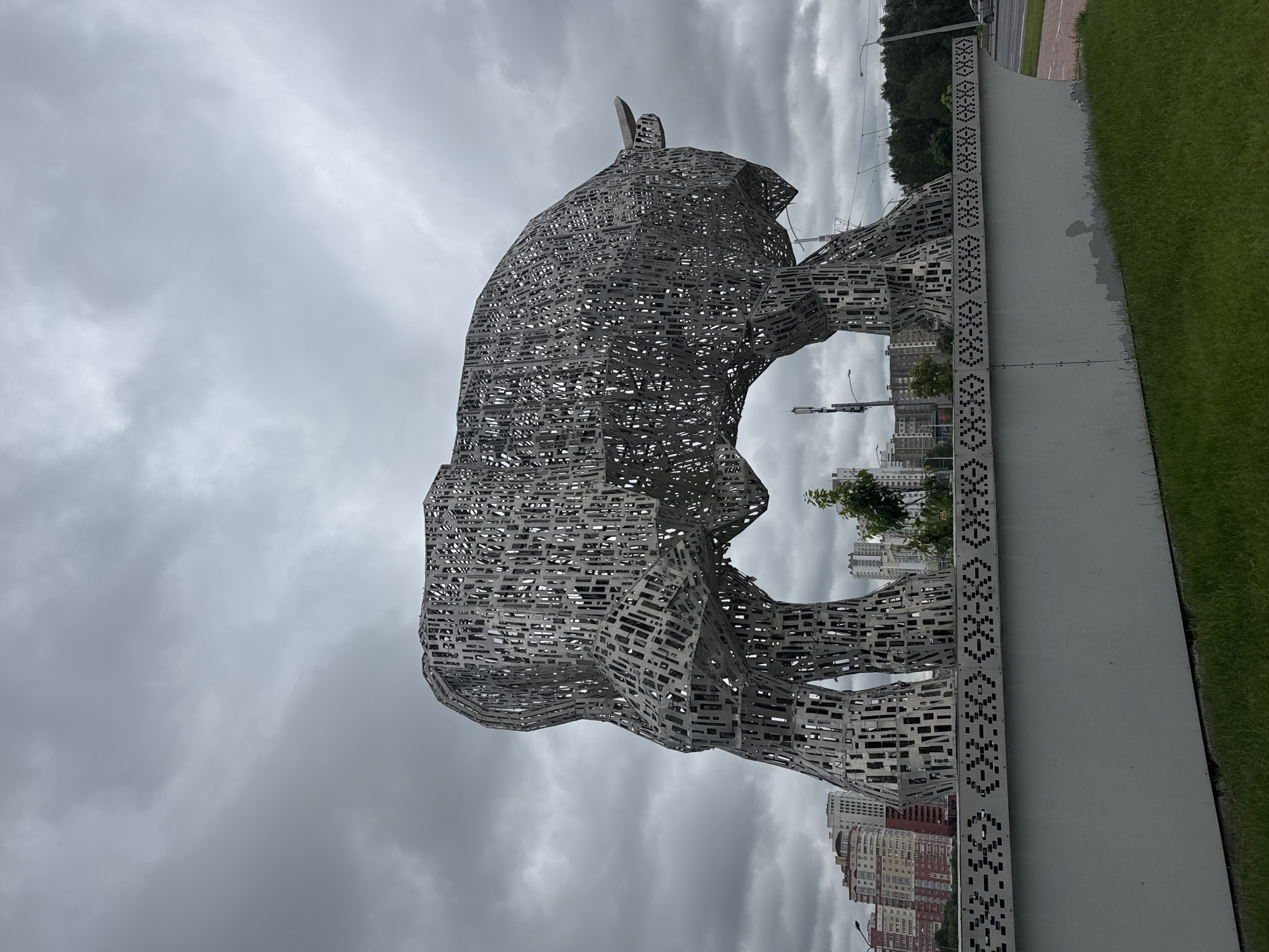 This image shows a large, abstract sculpture of a bull, made of metal, in the city of Minsk, Belarus. The sculpture is located near an urban area, with several apartment buildings visible in the background. The sky is overcast with clouds. The bull sculpture is placed on a concrete base that has a decorative pattern. The sculpture itself is composed of many small rectangular and square pieces of metal, creating a hollow, grid-like effect. The bull is depicted in a walking or standing pose, with its head facing to the right. There are no people visible in the image. The overall scene suggests an outdoor public space in a city.