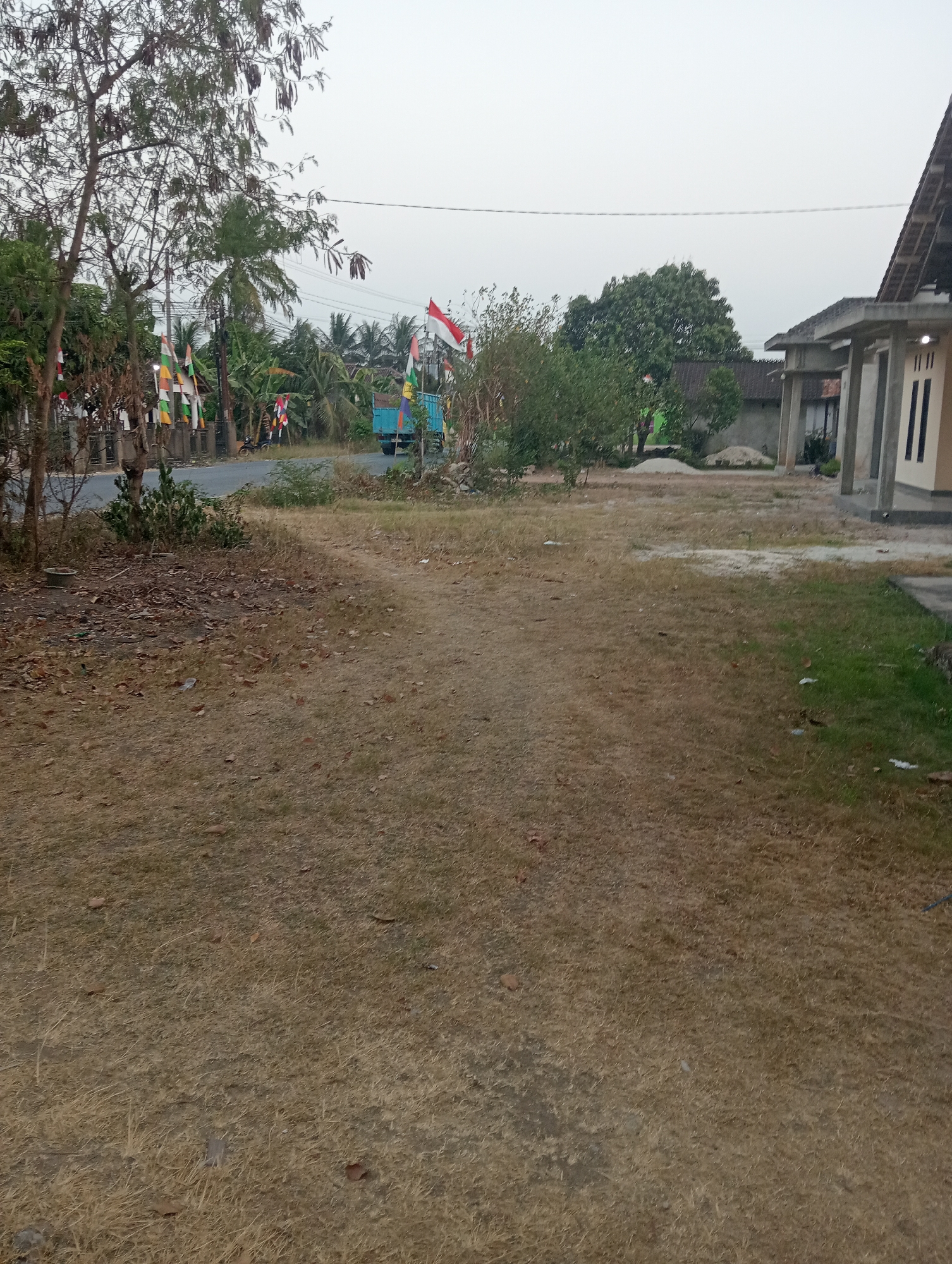 The image depicts an outdoor scene in Pringsewu, Indonesia, likely a rural or semi-urban area. In the foreground, a dirt path cuts through dry, brown grass. To the left, a road runs alongside a fence adorned with numerous colorful flags, possibly for a local celebration or national holiday. Trees, some with dry seed pods, are also present on the left. Further down the road, a blue truck is visible, also decorated with flags. On the right side of the image, a portion of a building under construction is visible, with concrete pillars and a roof structure. The background consists of trees, more buildings, and a hazy, overcast sky. The overall impression is one of a quiet, possibly developing area.