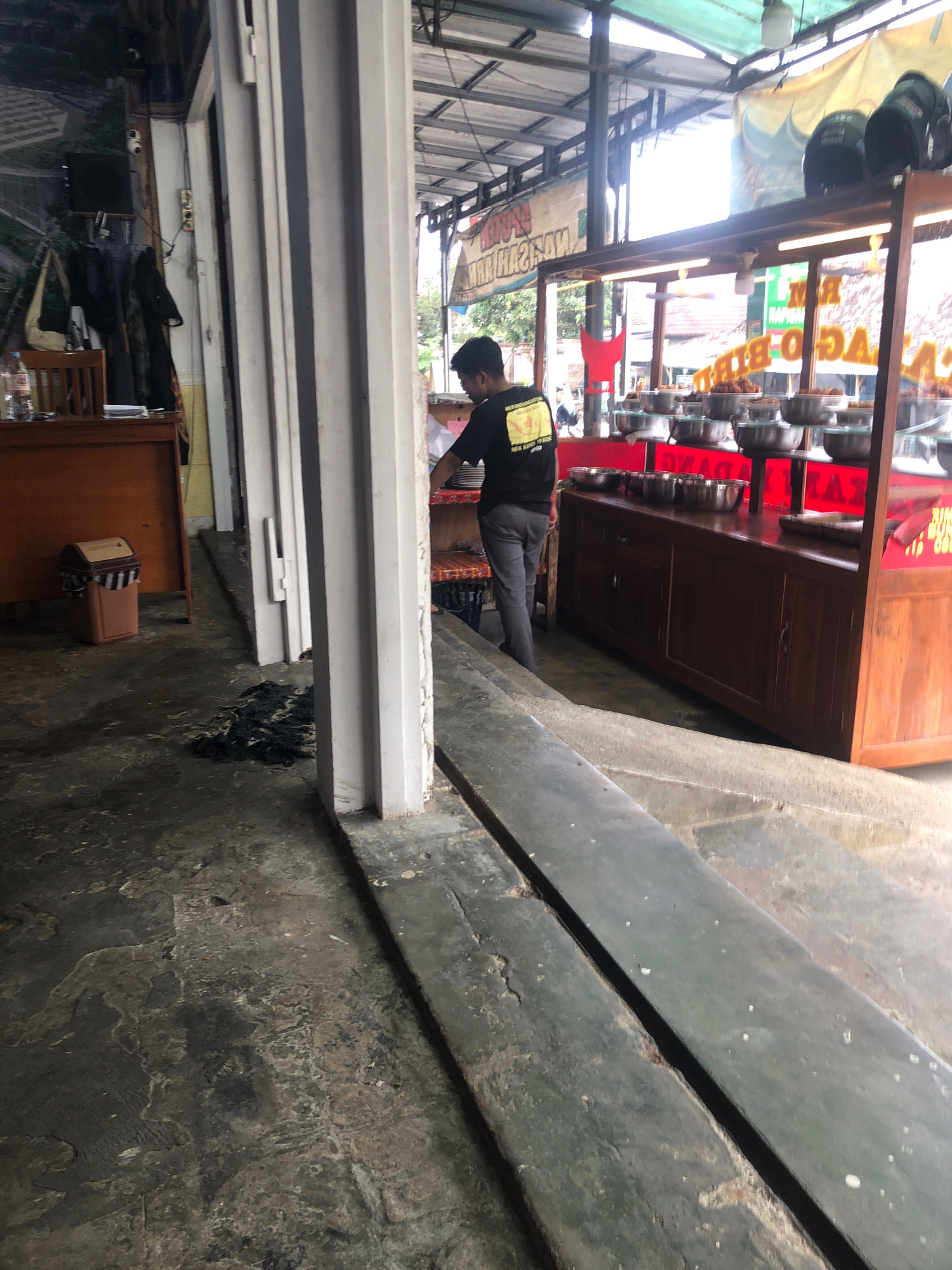 A man is standing at a food stall in Mendolo Lor, Indonesia, near a display case filled with food. The food stall has a wooden counter and a glass display. There are metal bowls with food in them on shelves. The man is wearing a black t-shirt with a yellow logo and gray pants. He is facing away from the camera and appears to be serving food. The stall is under a metal roof with some banners hanging above. The ground in front of the stall is concrete. To the left of the stall, there is a wooden desk with a trash can.
