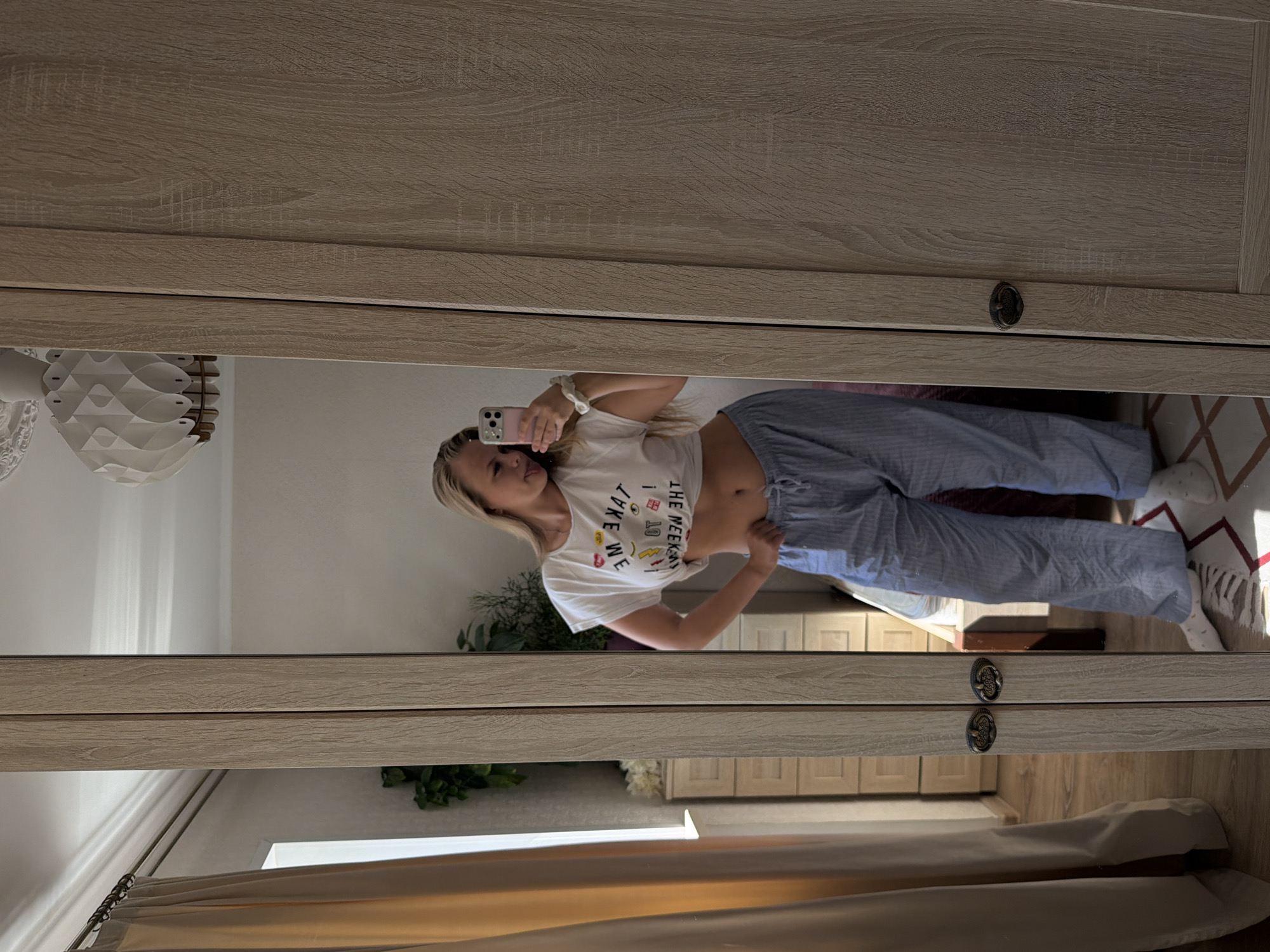 A woman with blonde hair is standing in front of a mirror, taking a selfie with her phone. She is wearing a white cropped t-shirt with a graphic print and blue and white striped pajama pants. Her left hand is on her hip, and she is sticking her tongue out at the camera. The mirror is framed by a wooden wardrobe with ornate handles. In the reflection, a cream-colored curtain and a dresser with drawers are visible. The overall environment suggests a bedroom. This scene is located in the city of Chaikovsky, Russia.