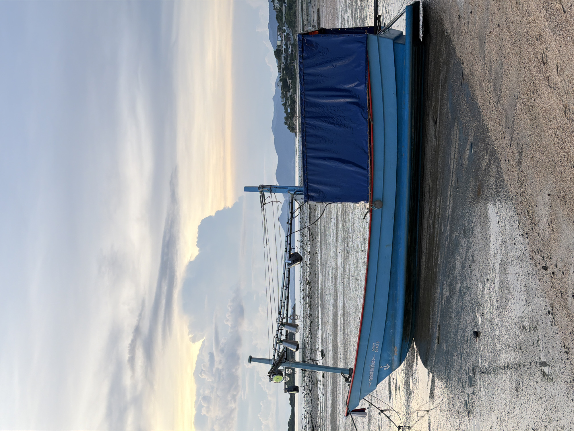 A light blue fishing boat with a red trim rests on a muddy, sandy shore. The boat is covered with a dark blue tarp on its right side. It is tethered by ropes and has an overhead structure with lights and bells. In the background, a calm body of water is visible, with a distant coastline featuring hills and scattered buildings. The sky above is cloudy with streaks of light, suggesting either sunrise or sunset. The scene is located in Baan Plai Laem, Thailand, and depicts a tranquil, coastal environment during a period of low tide. No people are present in the image.