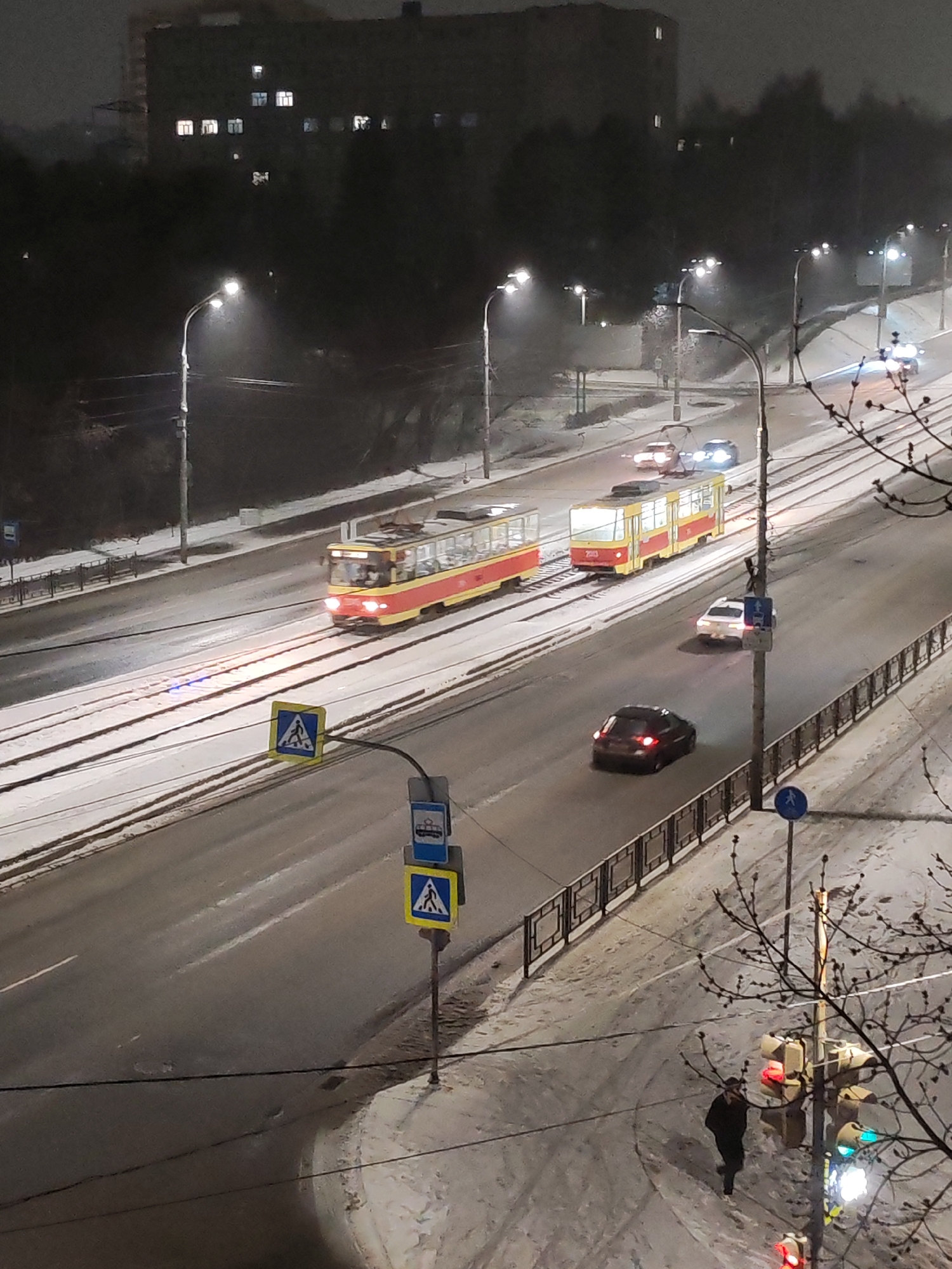 The image depicts a nighttime scene in Izhevsk, Russia, during winter. Two red and yellow trams are traveling on rails that run parallel to a road with multiple lanes of traffic. Cars are driving on the road, including a dark-colored sedan, a white car, and others further down the road. Streetlights illuminate the scene. On the right side of the image, there is a railing along the road and a snowy sidewalk with a pedestrian walking near a traffic light. Street signs are visible near a pedestrian crossing. A large building can be seen in the background.