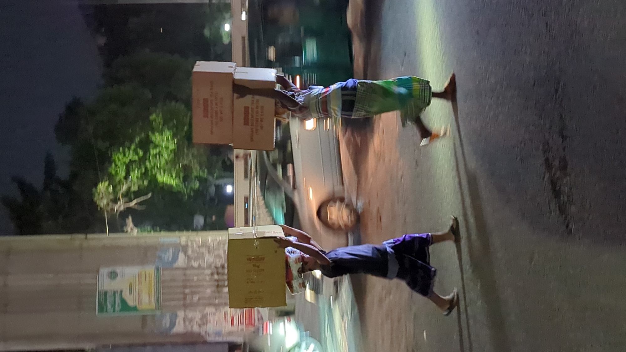 Two men are carrying boxes on their heads. They are walking on a paved road next to a building. A van is in the background. The scene is lit by artificial lights, appearing to be at night. The location is likely Dhaka, Bangladesh, inferred from the subjects' attire and the surrounding environment.