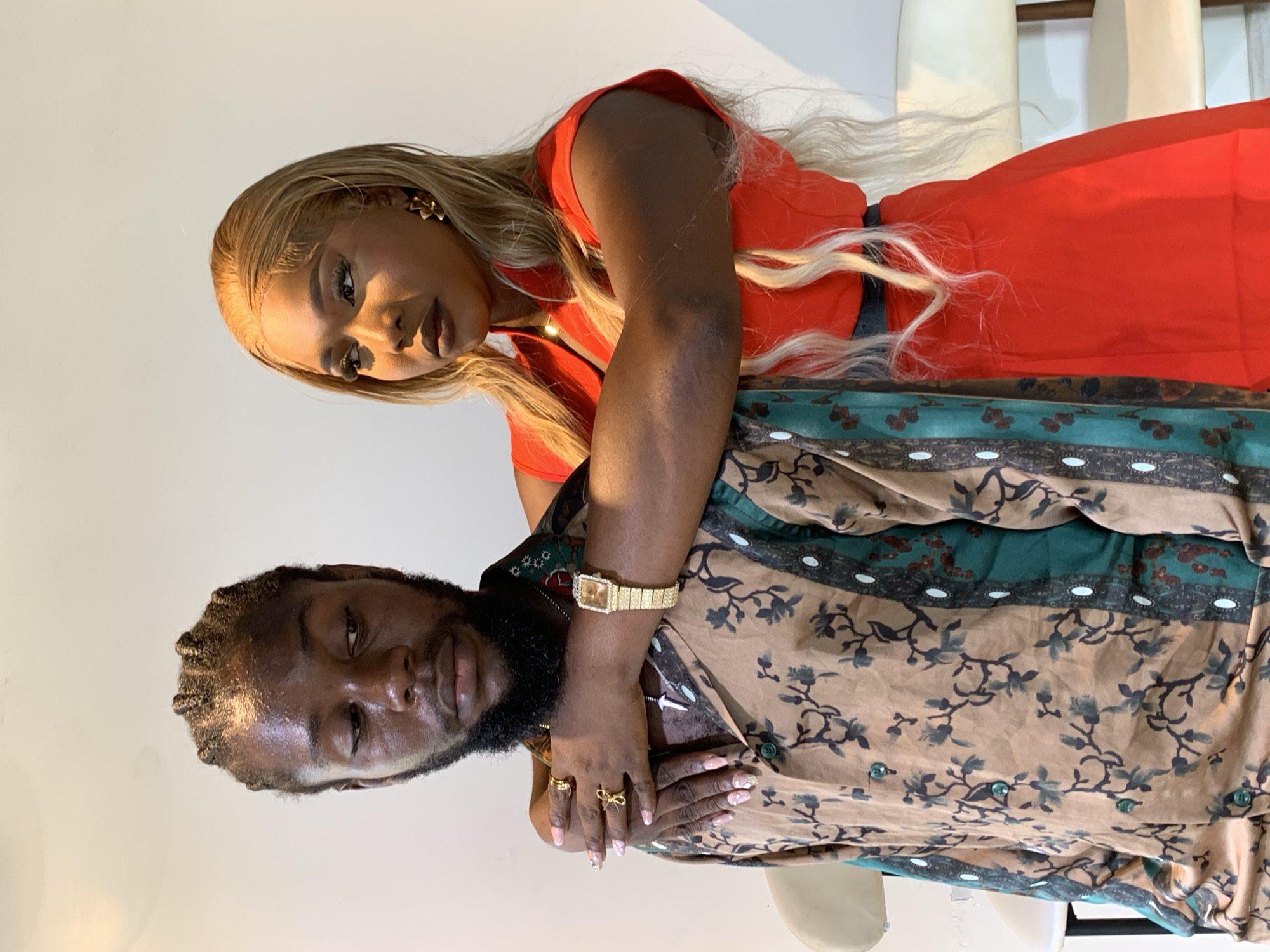 The image depicts two individuals, a man and a woman, posing for a photograph against a plain white background. The man, positioned on the left, has his head turned downwards and his eyes are closed. He has braided hair and a beard, and is wearing a patterned button-down shirt with floral motifs in brown and green. He is adorned with several gold rings and a silver pendant necklace.
The woman, on the right, has her arm around the man's shoulders, her hand resting on his chest. She has long, blonde hair and is wearing a bright red, sleeveless dress with a high collar. She is also wearing gold earrings and a delicate necklace. Her gaze is directed to her left, away from the camera. The lighting in the scene is bright, casting subtle shadows on the individuals and the background.
The scene does not provide any specific visual cues to indicate a location within the city of Ikorodu, Nigeria. The environment is minimalist, consisting only of the two subjects and the backdrop.