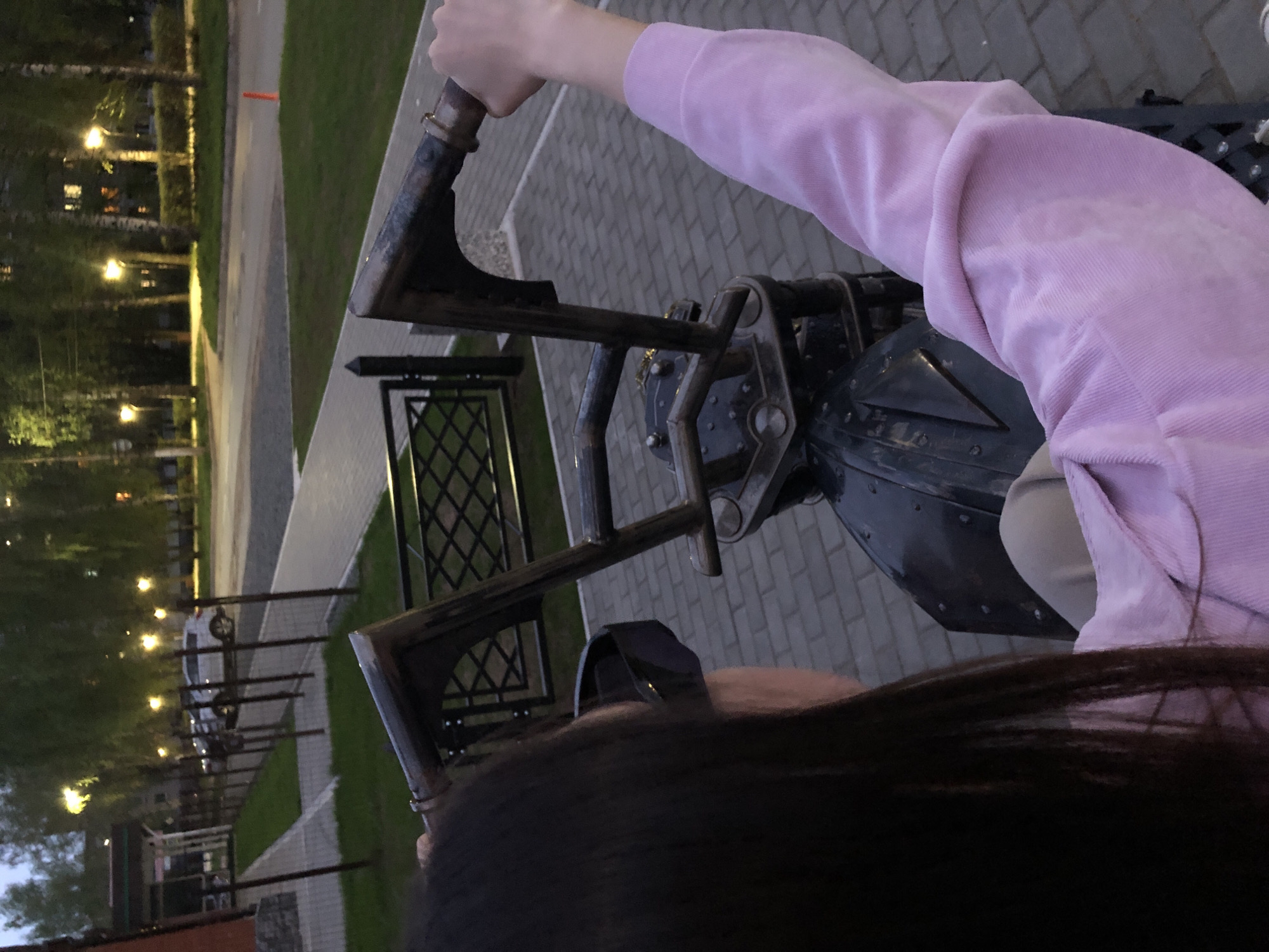 The image shows a person riding what appears to be an elaborate, steampunk-style mechanical sculpture or device. The sculpture is constructed of dark metal and features prominent rivets and structural elements. The person is wearing a light pink sweatshirt and has dark hair. The background suggests an outdoor setting, possibly a park or public area, with trees and decorative streetlights visible. The scene is in Chaikovsky, Russia. There are brick sidewalks and metal railings in the background.
