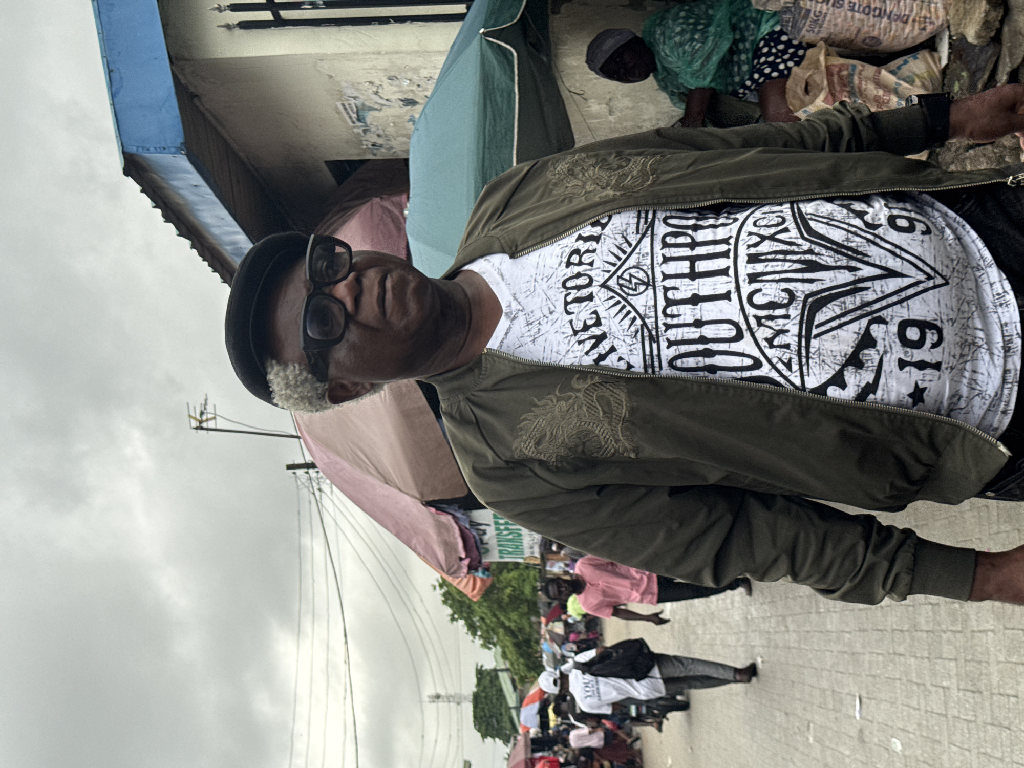 The image shows a man wearing a black hat, eyeglasses, and a green jacket over a white t-shirt with a graphic print. The man is positioned in front of what appears to be a street scene. To his right, there is a woman wearing a head covering seated near bags. Behind the man, there is a busy street with people walking and riding motorcycles. The environment suggests a marketplace or commercial area with buildings and overhead structures like umbrellas or awnings. The sign that reads "TRANSFER" suggests a place for monetary transactions. Based on the style of dress, the architecture, and the presence of "Dangote Sugar" sacks, the location may be Lagos, Nigeria. The day seems overcast, judging from the cloudy sky.