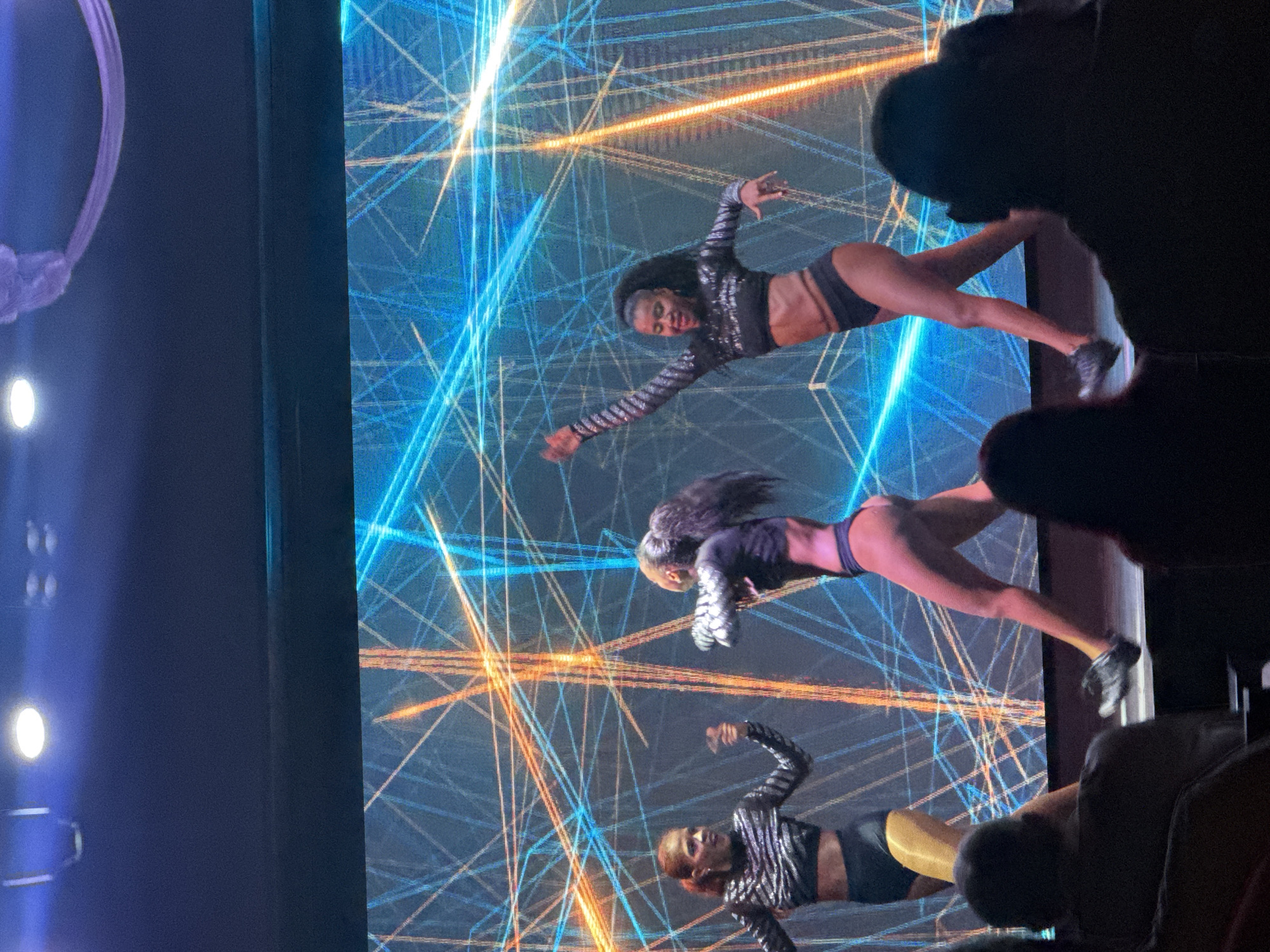 A medium shot captures three young women performing on a stage in front of a large LED screen displaying a dynamic, geometric pattern of blue and orange lines. The women are dressed in matching black, sequined crop tops and black bottoms. The woman on the left has auburn hair tied back and is looking forward, her left arm raised. The woman in the center has dark, curly hair pulled into a high ponytail and is facing away from the camera, her arms bent. The woman on the right has dark, curly hair also in a high ponytail and is facing the camera with her right arm extended upwards and her left arm bent. The foreground shows the silhouettes of audience members seated in darkness. The lighting is stage lighting, with spotlights visible at the top of the frame. The overall atmosphere suggests a performance or entertainment event. The location is identified as Manavgat, Turkey.