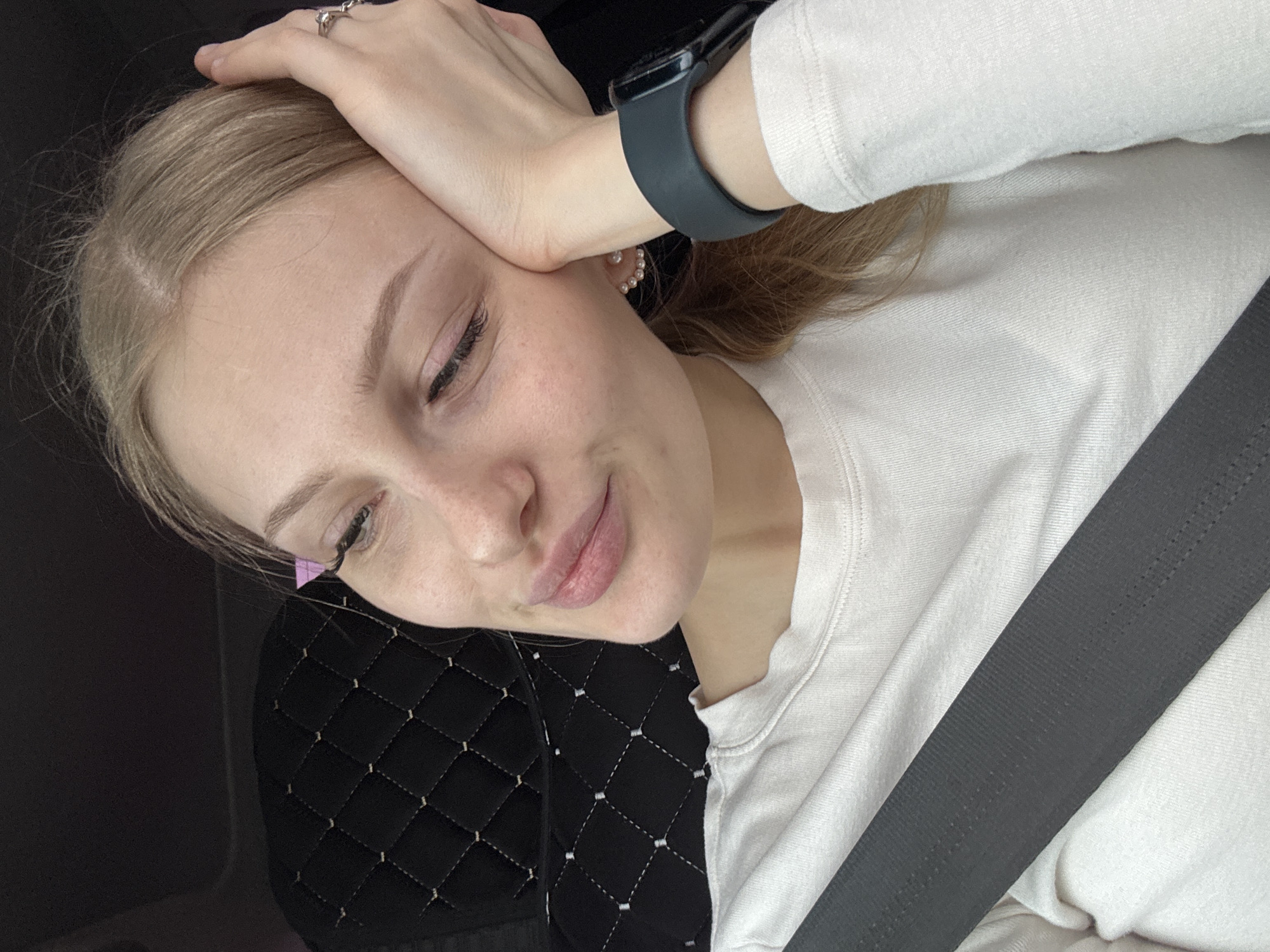 A young, fair-skinned woman with blonde hair is seated in what appears to be a vehicle. She is wearing a white long-sleeved shirt and a dark-colored smartwatch on her left wrist. A seatbelt is fastened across her torso. Her left hand is resting on her head, and she has a ring on one of her fingers. The car seat has a black quilted pattern. There's no indication of the location being Perm, Russia in the image.