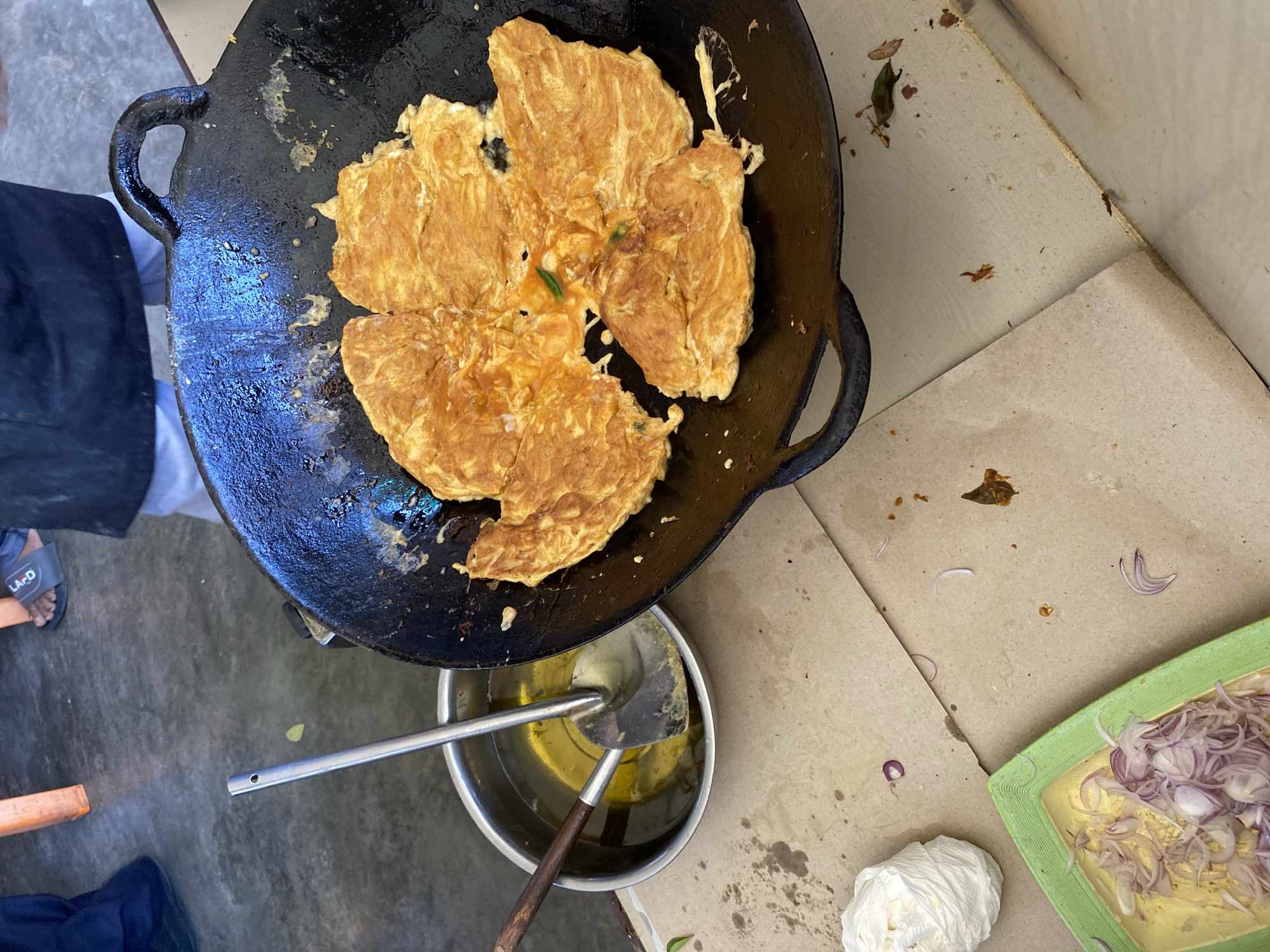 An overhead, slightly angled shot captures a street food cooking scene in Banda Aceh, Indonesia. The primary subject is a large, black wok filled with a golden-brown, flower-shaped omelet, its edges slightly crisped. The omelet appears to have been cooked in sections, radiating from the center. To the left of the wok, a stainless steel bowl contains a generous amount of cooking oil, with a metal spatula and a wooden-handled ladle submerged within it. The cooking station is set up on a textured, light brown surface, likely a table or counter covered with paper. Scattered on this surface are remnants of cooking, including small green leaves, oil splatters, and a crumpled white paper towel. In the lower left corner, a green plastic tray holds thinly sliced red onions. In the background, a person's legs are visible, wearing dark pants and black slides with the word "LAJO" printed on them, along with the edge of an orange plastic chair. The flooring appears to be concrete.