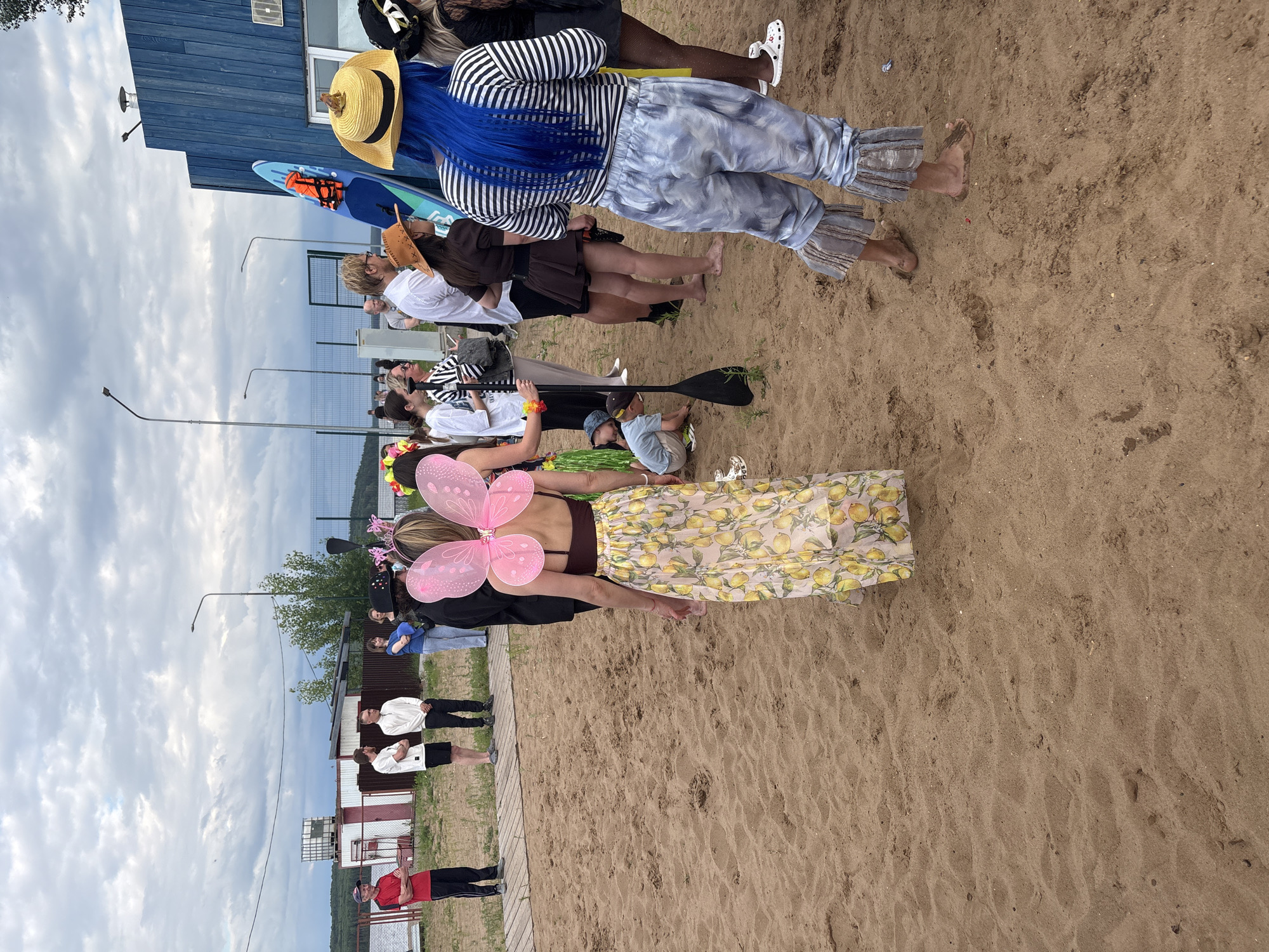This media file, taken in Chaikovsky, Russia, captures a lively outdoor gathering on a sandy beach. A group of people are assembled, some in costumes, suggesting a playful event or party. One person stands out with bright blue hair and a straw hat, wearing a striped top and tie-dye pants. Another individual is dressed with pink butterfly wings and a yellow skirt adorned with lemons, adding a whimsical touch to the scene. Other people in the background are dressed casually, some in t-shirts and shorts, while others are in more elaborate outfits.
Objects scattered around include a blue paddleboard with an orange life vest, and a paddle. The setting appears to be near a wooden building with a blue facade and a white window frame. The ground is sandy with some footprints. The sky is overcast with clouds, indicating a day that is not intensely sunny. The overall mood seems relaxed and festive, with people engaged in conversation and enjoying the outdoor environment. The presence of beach attire and recreational equipment suggests a summer or warm-weather event. There is no visible text in the image.