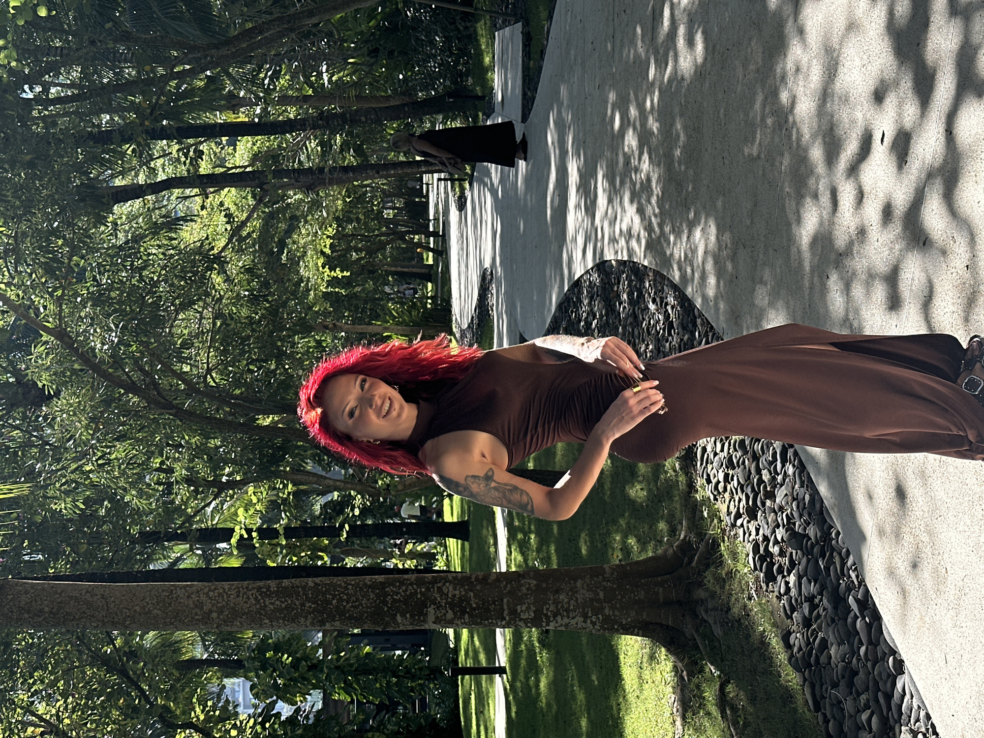 The image depicts a woman with red hair standing outdoors, likely in a tropical or subtropical environment. She is wearing a brown, long dress. She is posing, smiling, and looking directly at the camera. The background consists of lush greenery with trees. A path or walkway constructed of concrete and lined with black pebbles is visible to her right. In the background along the path another person is visible. Given the prompt's location reference, it is likely that the image was taken in Kuta Selatan, Indonesia, within a park or garden featuring a paved pathway.